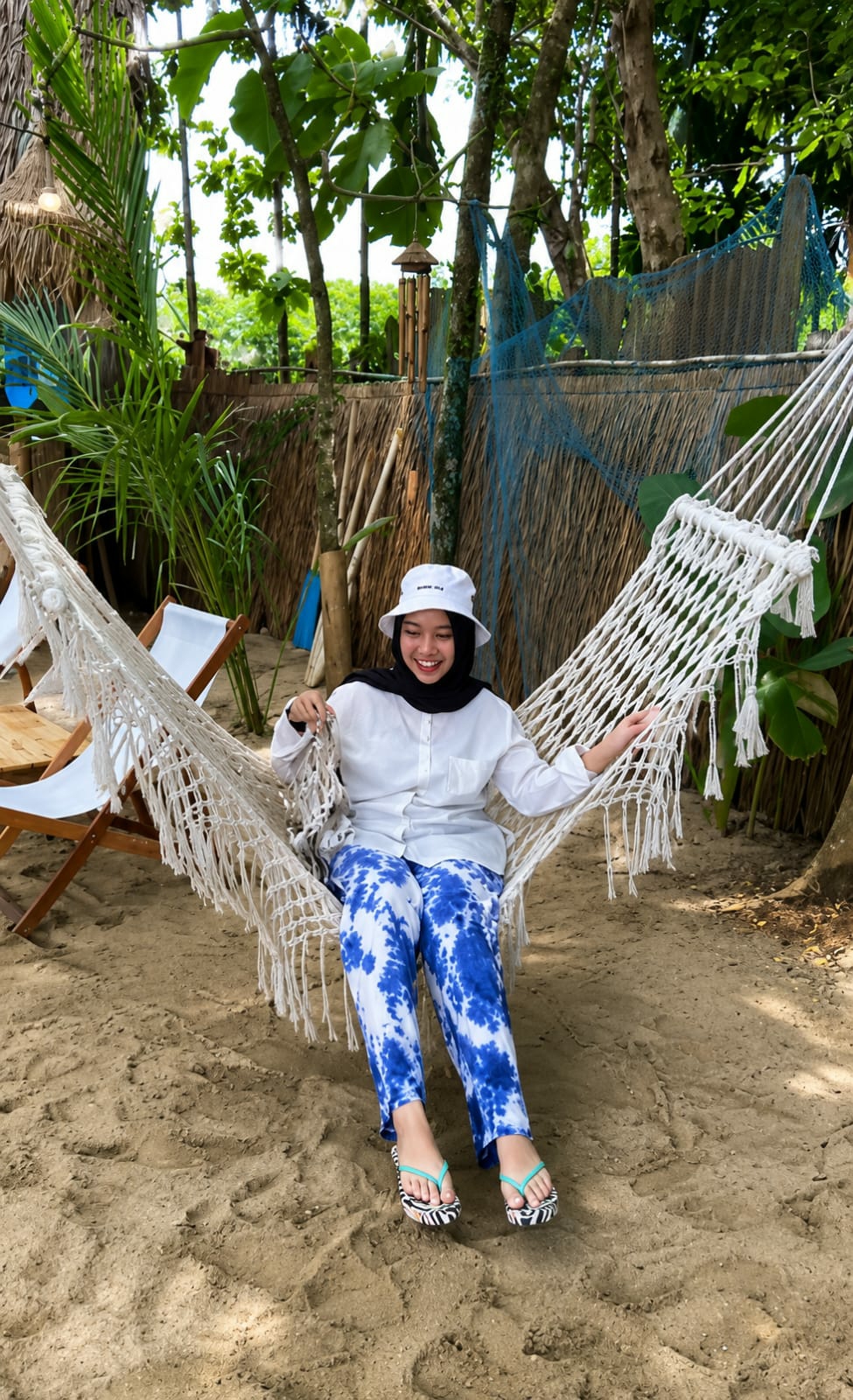 A young woman wearing a white bucket hat, black hijab, white button-up shirt, and blue and white tie-dye pants is sitting in a white macrame hammock on a sandy beach. She is smiling and looking at the camera, with her left hand resting on the hammock. Her feet are resting on the sand, and she is wearing patterned flip-flops. Behind her is a backdrop of palm trees, tropical foliage, and a thatched-roof structure. A blue net is draped over a fence made of bamboo and thatch. The scene appears to be an outdoor relaxation area at a beach resort. The city of Medan, Indonesia is characterized by its tropical climate and numerous beaches, which are popular tourist destinations.