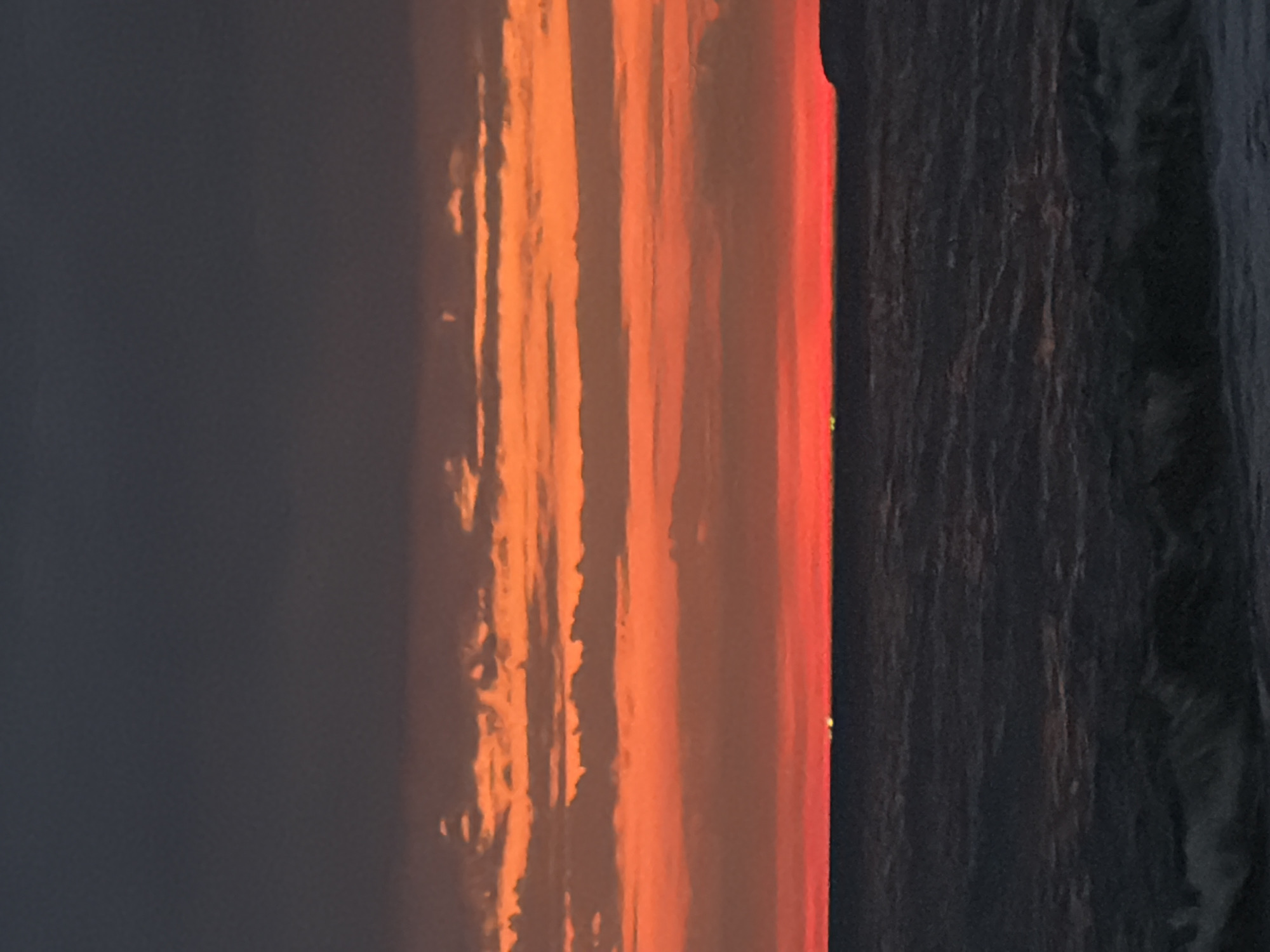 This image depicts a sunset over the Black Sea, possibly near Sochi, Russia. The sky is a vibrant gradient of dark gray at the top transitioning through various shades of orange and red towards the horizon. Horizontal cloud formations are visible in the orange bands. The sea below is dark and choppy, with visible waves. On the horizon, there are several small, bright green lights scattered across the boundary between the sea and the sky, and a faint landmass is visible on the far right. The overall impression is of a dramatic and colorful natural scene at dusk.