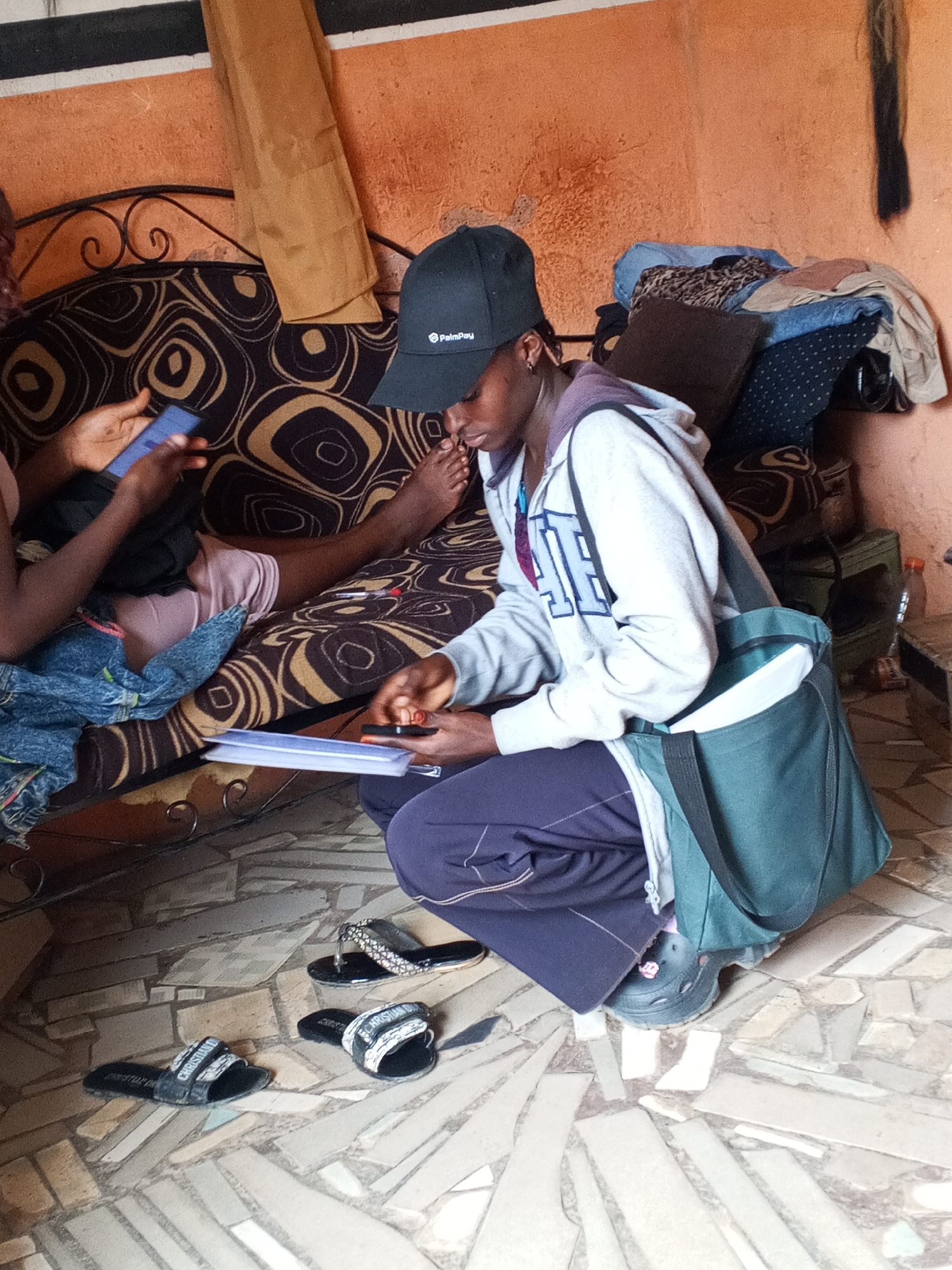 The image captures a scene indoors, likely in a home in Gwong, Nigeria. The setting appears modest. The walls are textured and painted in a light orange hue. The floor is tiled with irregular, light-colored tiles.
Two individuals are present. One is seated on a couch covered in a black and brown circular pattern fabric, looking at a smartphone. The second individual, wearing a PalmPay-branded baseball cap, a grey hooded sweatshirt with a collegiate-style emblem, and dark-colored pants, is kneeling on the floor. They are holding a clipboard with papers and appear to be focused on it. They have a green shoulder bag. A pair of sandals sits near the kneeling person.
The couch has items piled on it including clothing and a pillow. A water bottle is visible on the table next to the couch. A piece of dark hair is on the wall to the right.