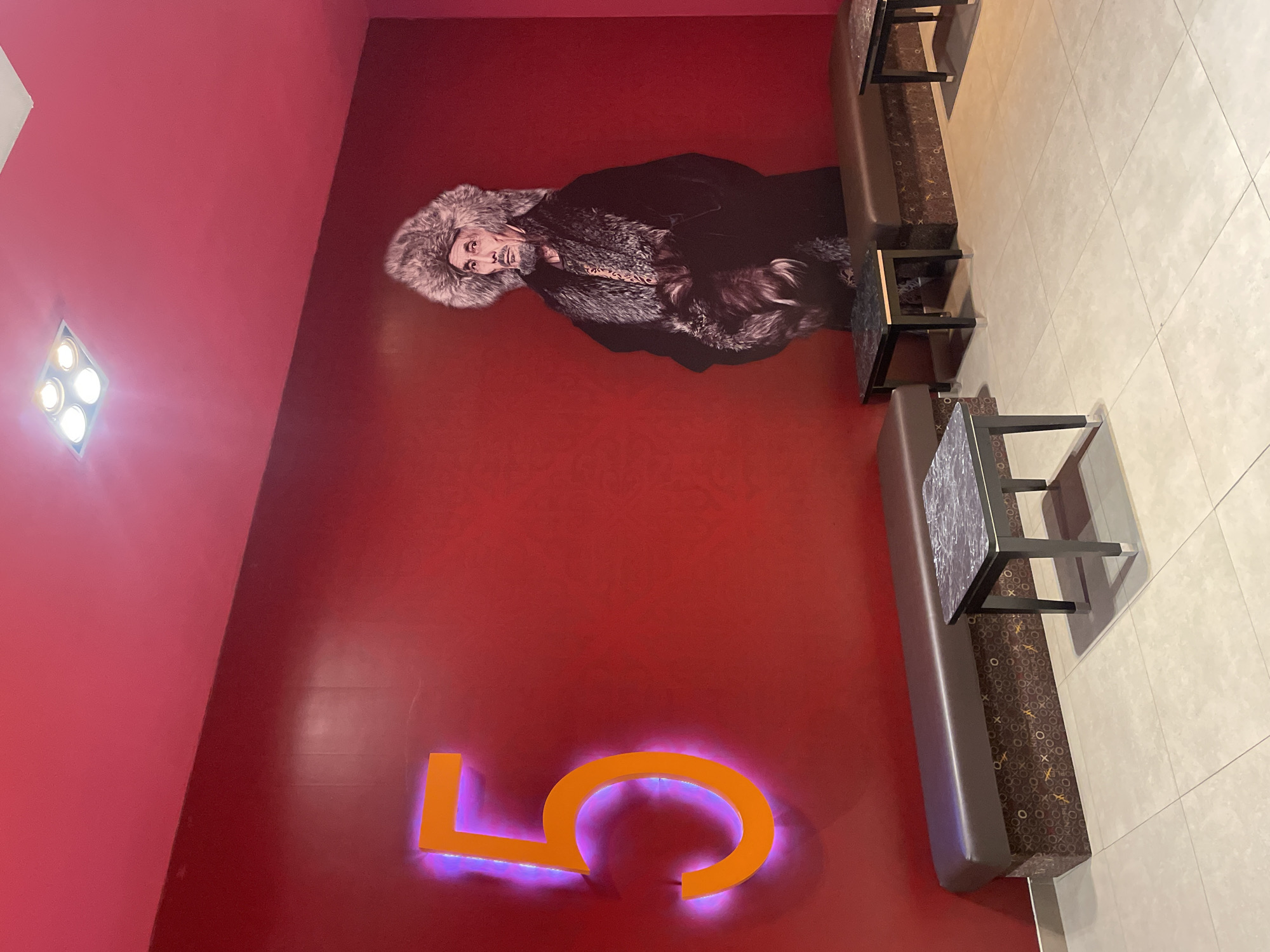 The image depicts an interior space with a vibrant red wall. On the left side of the wall, a large, illuminated orange number "5" is prominently displayed. To the right, a life-sized cutout of a man wearing a fur hat and a fur-lined coat stands against the red wall. In front of the wall, there are several brown leather benches with patterned bases and small, dark-colored tables with marble-like tops. The floor is tiled in a light beige color. The scene appears to be a waiting area or lounge, possibly within a cinema or entertainment venue. The image is taken from a frontal perspective, at approximately eye level. The lighting is bright, with ceiling lights visible in the top portion of the image. Based on the prominent "5" and the style of the cutout, this location is likely "KinoPark 5," a cinema complex located in Almaty, Kazakhstan.