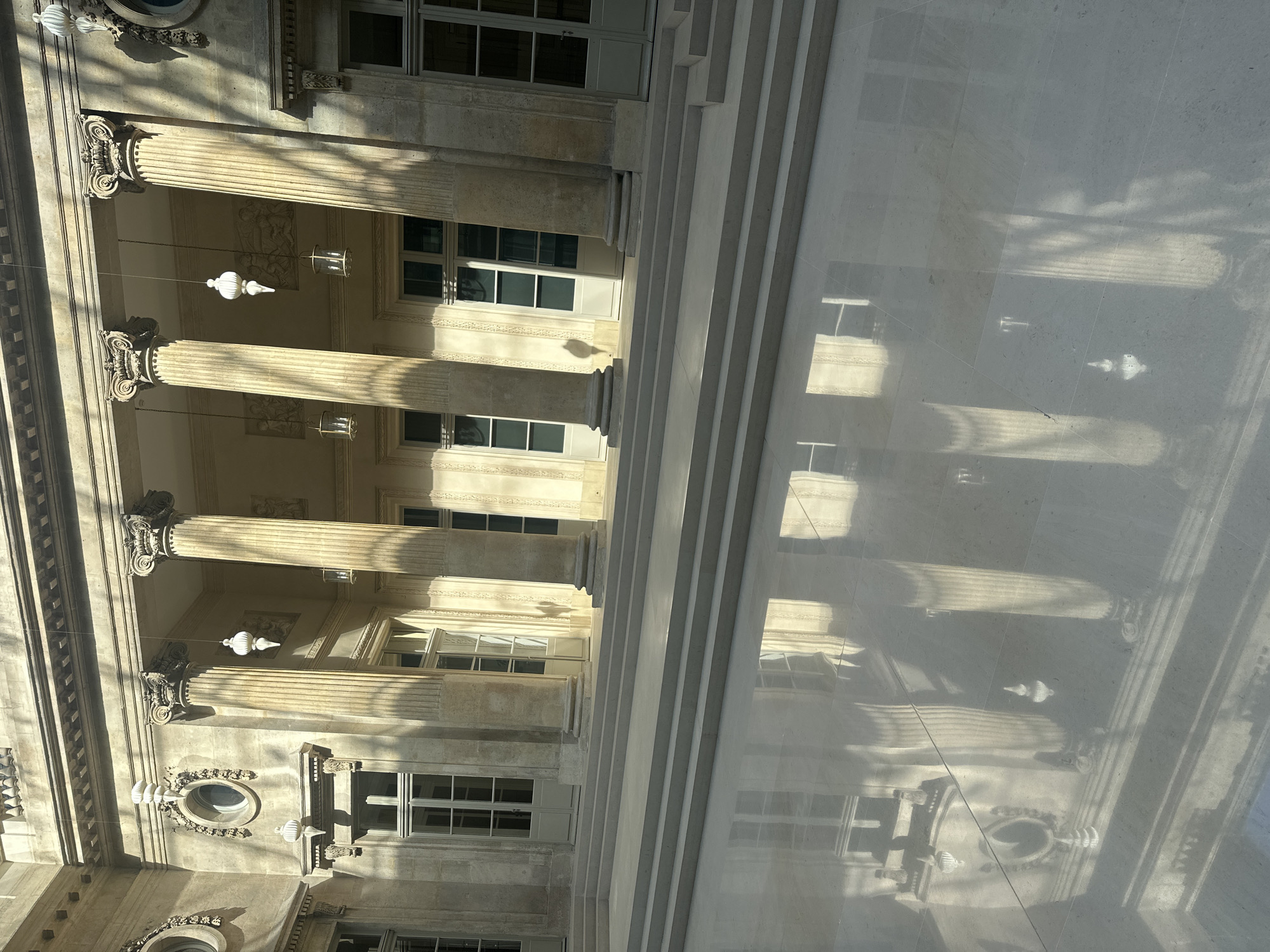 The image shows a building with classical architecture, possibly a museum or government building, in Paris, France. The building has a colonnade with decorative Corinthian columns and numerous windows. The light suggests it is daytime, and shadows are cast on the marble floor, reflecting parts of the building. There are no people visible. The architectural style, window design, and material suggest the location is within the city of Paris, France.