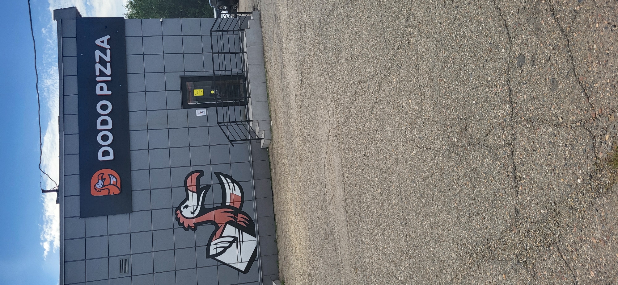The image shows a building with a sign that reads "DODO PIZZA". There is a mural of a dodo bird on the side of the building. The building appears to be made of metal panels and has a glass door with a railing and stairs leading up to it. The area in front of the building is paved with asphalt. The sky is blue with some clouds.