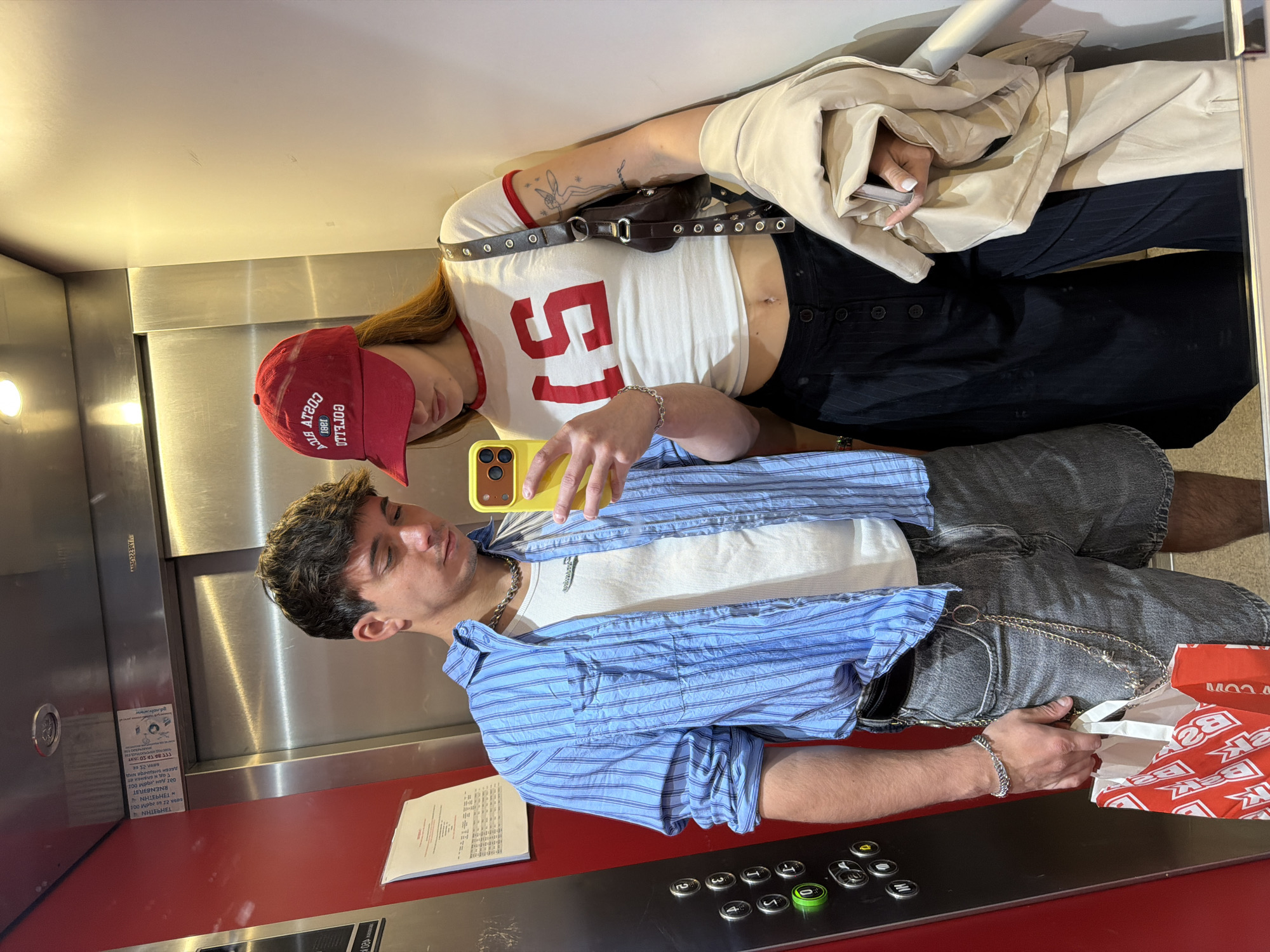 The image shows a couple in an elevator taking a selfie in front of a mirrored wall. The woman has long brown hair, is wearing a red baseball cap, a white cropped shirt with the number 51 in red, and black pants. A light-colored coat is draped over her shoulder. She wears a black belt with silver eyelets. The man has short brown hair and wears a blue striped shirt over a white t-shirt, and gray shorts. A silver chain is visible on his shorts. He holds a yellow smartphone. A white document is visible on the elevator's control panel, which is red and silver. The control panel has round buttons. A paper bag with branding is visible in his hands. The location is an elevator interior, likely in Sofia, Bulgaria, based on text on the document.