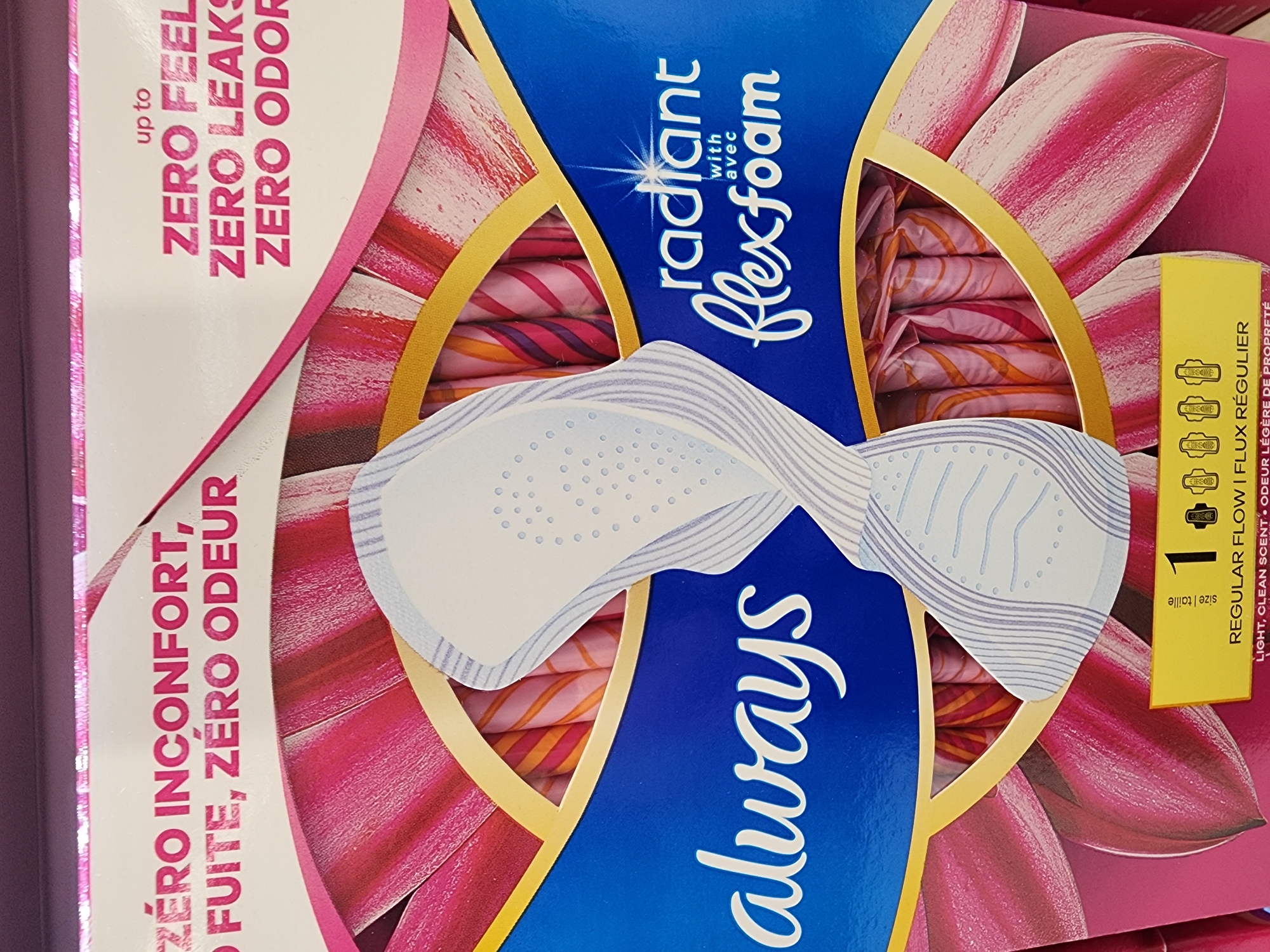 This is a close-up, eye-level shot of a package of Always Radiant FlexFoam pads. The packaging features a dark blue background with a bright pink floral design. The "always" logo is prominently displayed in white script across the bottom left of the package. Above the logo, in white text, it reads "ZÉRO INCONFONT, O FUITE, ZÉRO ODEUR" which translates to "ZERO DISCOMFORT, ZERO LEAKAGE, ZERO ODOR." To the right, in pink lettering, it says "up to ZERO FEEL," "ZERO LEAKS," and "ZERO ODOR." A stylized representation of a pad is shown in the center of the packaging. In the bottom right corner, a yellow label reads "1" with icons representing pads, and "REGULAR FLOW I FLUX RÉGULIER." This product is likely for sale in a retail environment. The location of Camp Hill, United States of America, is not discernible from the image itself, as it is a product shot and not a depiction of a specific location.