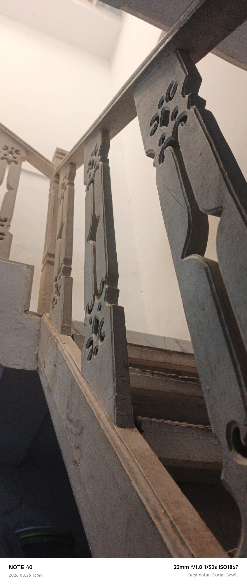The image shows a close-up view of an old wooden staircase with intricate carvings on the banisters. The staircase appears weathered and worn, suggesting it is an older structure. The lighting is dim, casting shadows that highlight the texture and details of the wood. The background is a plain white wall, creating a contrast with the dark wood of the stairs. At the bottom of the image, there is text that reads "NOTE 40 2026.08.26 13:49" and "23mm f/1.8 1/50s ISO1867 Kecamatan Duren Sawit". This indicates that the photo was taken in Kecamatan Duren Sawit, which is a subdistrict within the city of East Jakarta, Indonesia. There are no people or animals visible in the image.