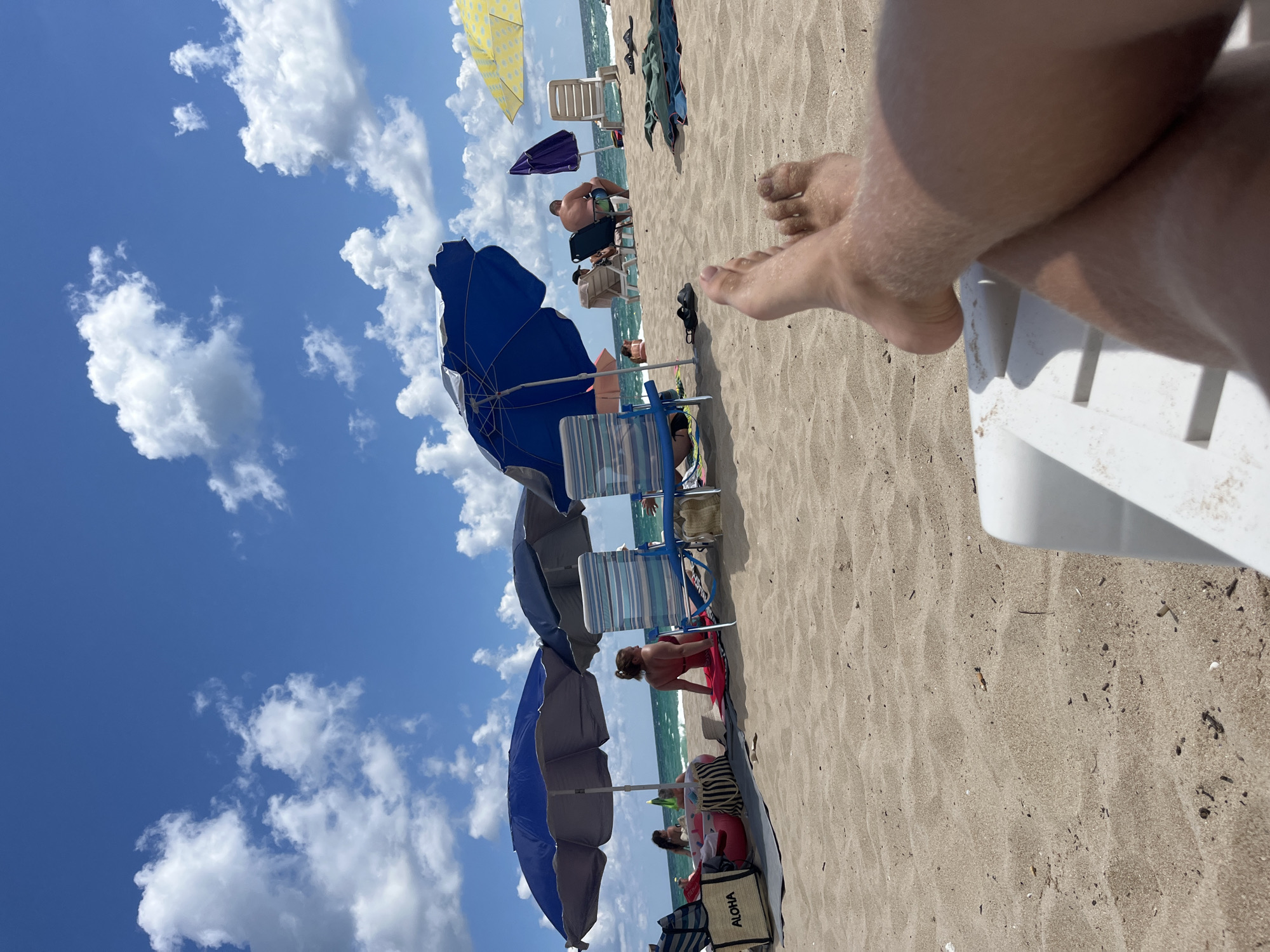 This image depicts a crowded beach scene with multiple beach umbrellas and lounge chairs set up on the sand. Several people are visible relaxing on the beach, some under umbrellas and others sunbathing. The ocean is visible in the background with waves and a blue sky with scattered clouds above. In the foreground, a pair of bare feet are resting on a white lounge chair. The image is taken from a low angle, looking out towards the beach and ocean. There is no information in the image to suggest this is in Primorsko, Bulgaria.