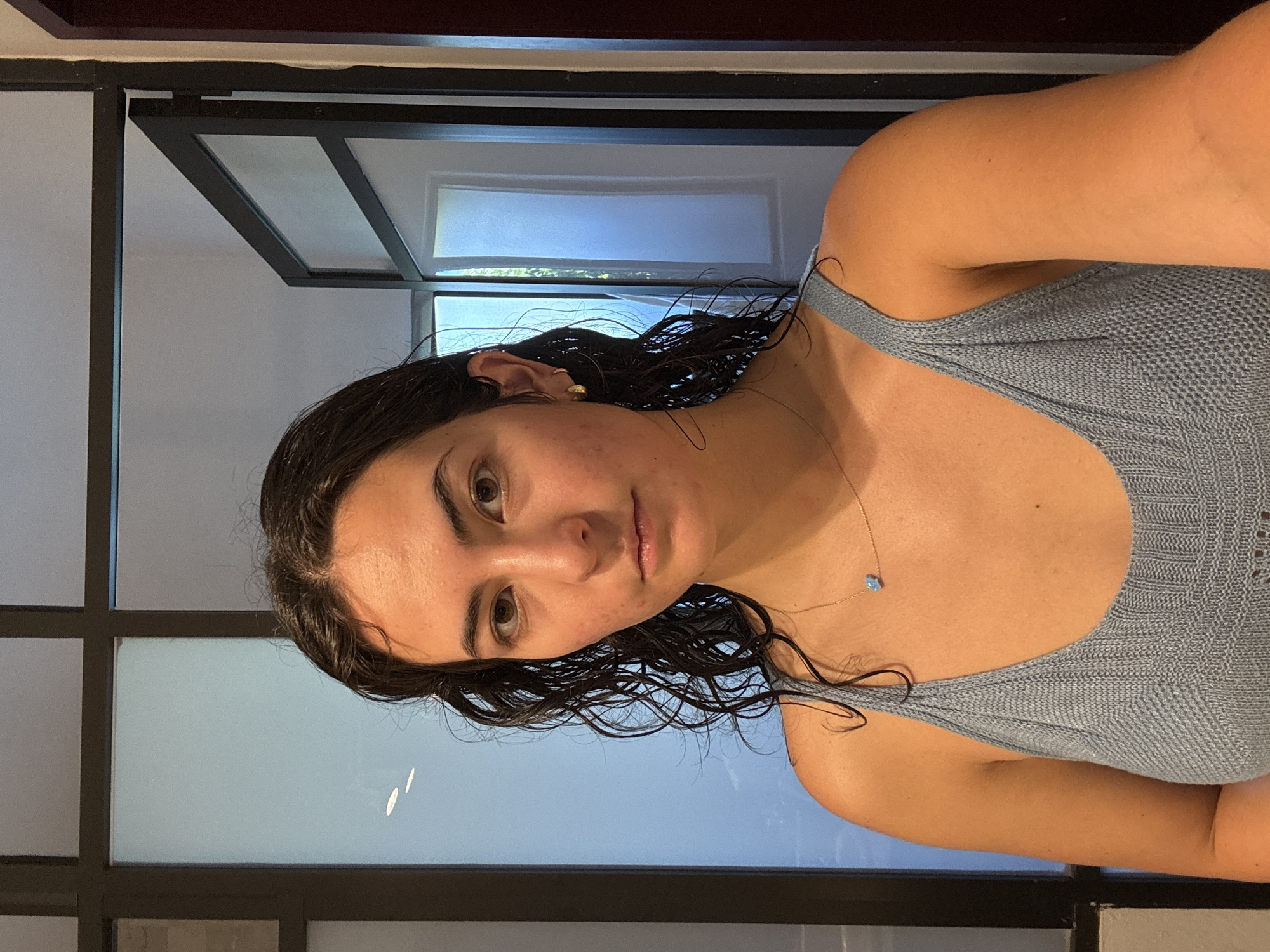 A young woman is depicted in a close-up, portrait-style image. She has dark, wet, wavy hair, dark brown eyes, and a neutral to slightly serious expression as she looks directly at the camera. Her skin tone is fair, with some visible redness and minor blemishes on her face. She wears a light blue or grey knitted tank top, featuring a distinct textured pattern. A delicate gold chain necklace with a small, light blue pendant is visible on her chest, and a small gold hoop earring is present in her left ear.
The background consists of dark-framed glass panels, which appear to be part of a door or large window, allowing diffused natural light to illuminate the scene. Reflections of light are visible on the glass surfaces. The image itself is oriented at a 90-degree angle, with the woman's head tilted to her right and her body extending downwards from that perspective. No text is visible in the media file. The wet appearance of her hair suggests she may have recently been in water or showered. The location context is Puebla, Mexico.