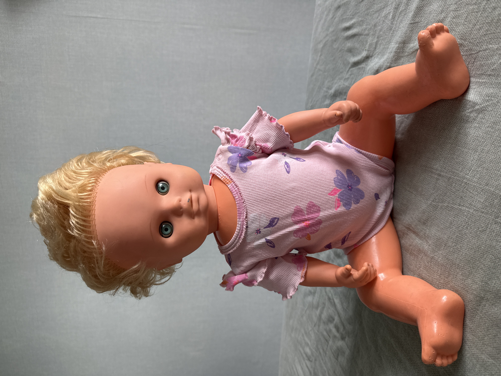 A vintage baby doll with blonde curly hair and blue eyes sits on a textured grey surface. The doll is wearing a pink one-piece outfit adorned with floral patterns in purple and pink. The doll's skin is a peachy-orange color, and its limbs are articulated. The background is a plain, light grey fabric. There are no people or animals present. The environment does not suggest a specific location in Moscow, Russia, or any other city.