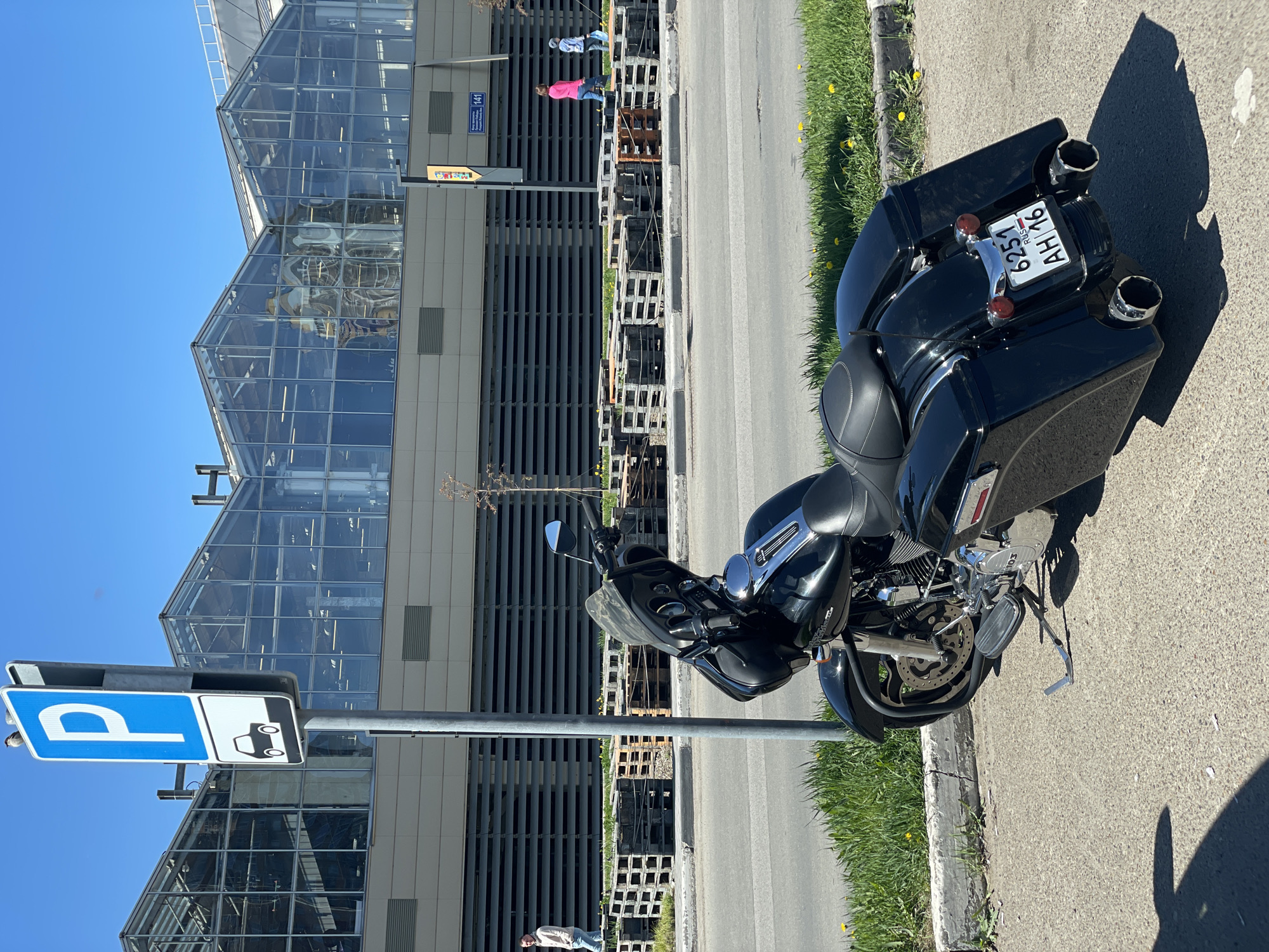 The image shows a black touring motorcycle parked on the sidewalk next to a parking sign with a "P" and a car symbol. The motorcycle has saddlebags and chrome accents. A Russian license plate reading "6251 AH 16 RUS" is visible. In the background, there is a modern building with a glass facade and gray horizontal metal slats. A stack of wooden pallets sits near the building. Two pedestrians, one in pink and one in blue, are walking along the sidewalk. The location is likely in Kazan, Russia based on the license plate. The weather appears sunny.