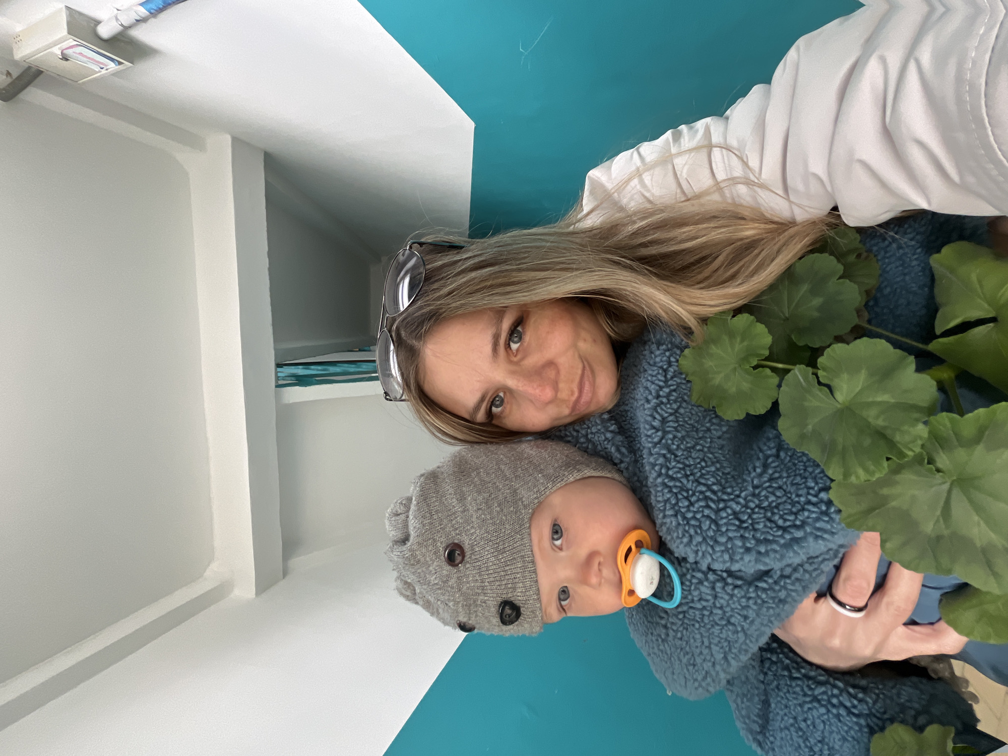 This image, taken in Chaikovsky, Russia, features a woman and a baby in an indoor setting. The woman, with long blonde hair and glasses resting on her head, smiles gently towards the camera. She is wearing a white long-sleeved top or jacket. In her arms, she holds a baby wearing a cozy blue, textured, fleecy outfit and a grey knitted hat decorated with dark buttons, resembling animal ears or eyes. The baby has a light orange and blue pacifier in its mouth and looks towards the camera with a neutral expression.
A vibrant green plant with large, rounded leaves is visible to the right of the subjects. The background consists of a bright indoor space with crisp white walls, contrasting sharply with a bold turquoise or teal colored wall or ceiling section. Architectural details such as a white beam or column, and a white light fixture on the ceiling, are visible in different parts of the room. The woman's left hand, visible in one of the detailed crops, sports a two-toned ring (black and white). The overall mood of the image is calm and affectionate. The time of day and external weather are not discernible, and no visible text is present.