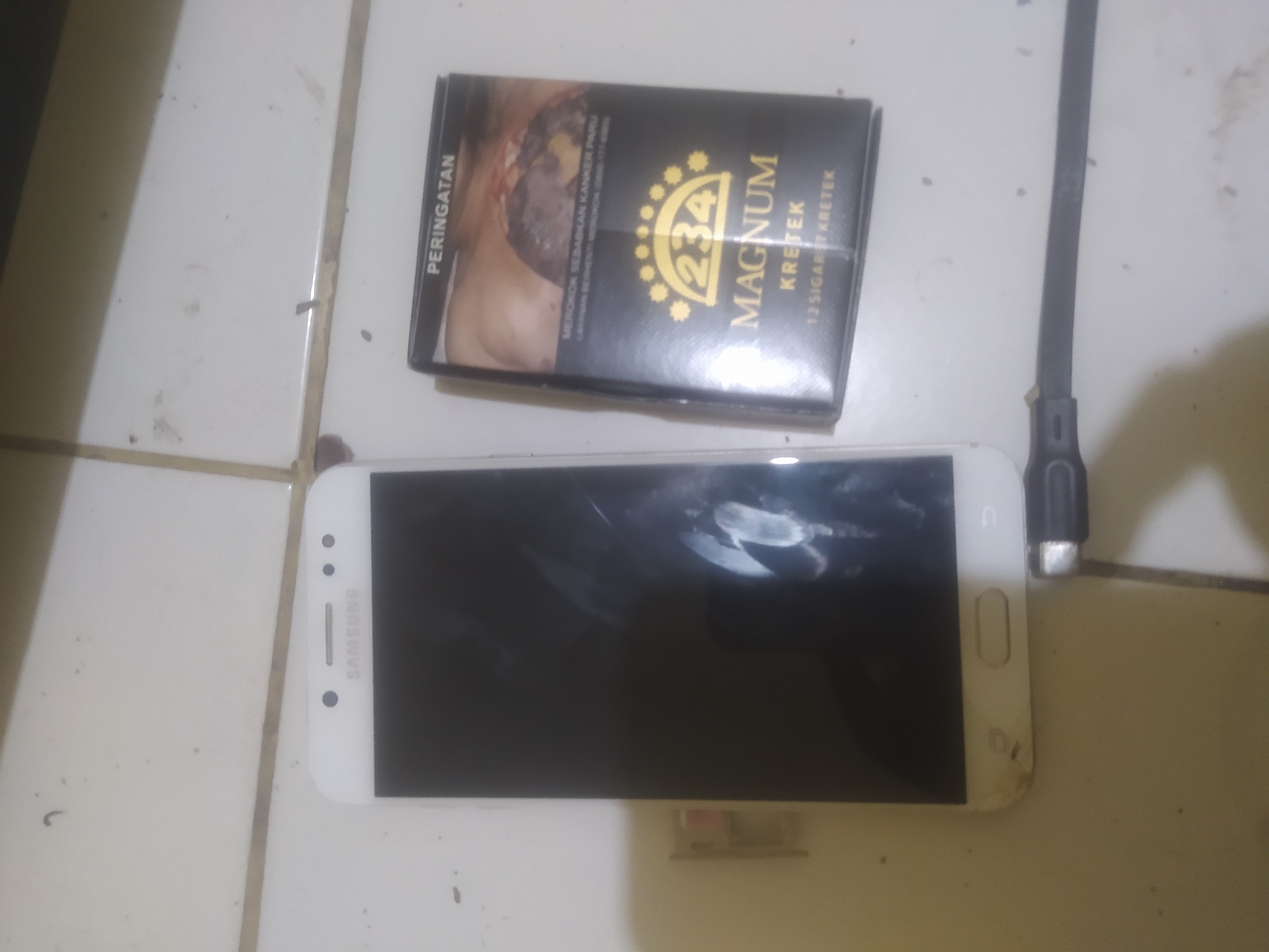 A white Samsung smartphone and a pack of Magnum Kretek cigarettes are on a tiled surface. The smartphone is positioned vertically to the left of the cigarettes, with its screen facing upwards. The cigarettes are in a black and gold pack, with text indicating "234 Magnum Kretek" and a warning label in Indonesian. A charging cable is partially visible in the foreground, connecting to the bottom of the smartphone. The tiled surface appears to be light-colored and is speckled with dirt and grime, suggesting an indoor environment. The overall lighting is dim. No people are visible in the image. The scene is static and does not depict any actions or interactions. The location is not explicitly identified, but the presence of Indonesian text on the cigarette pack suggests a connection to Indonesia. Without further information or context, it is impossible to pinpoint the exact location within Bekasi, Indonesia.