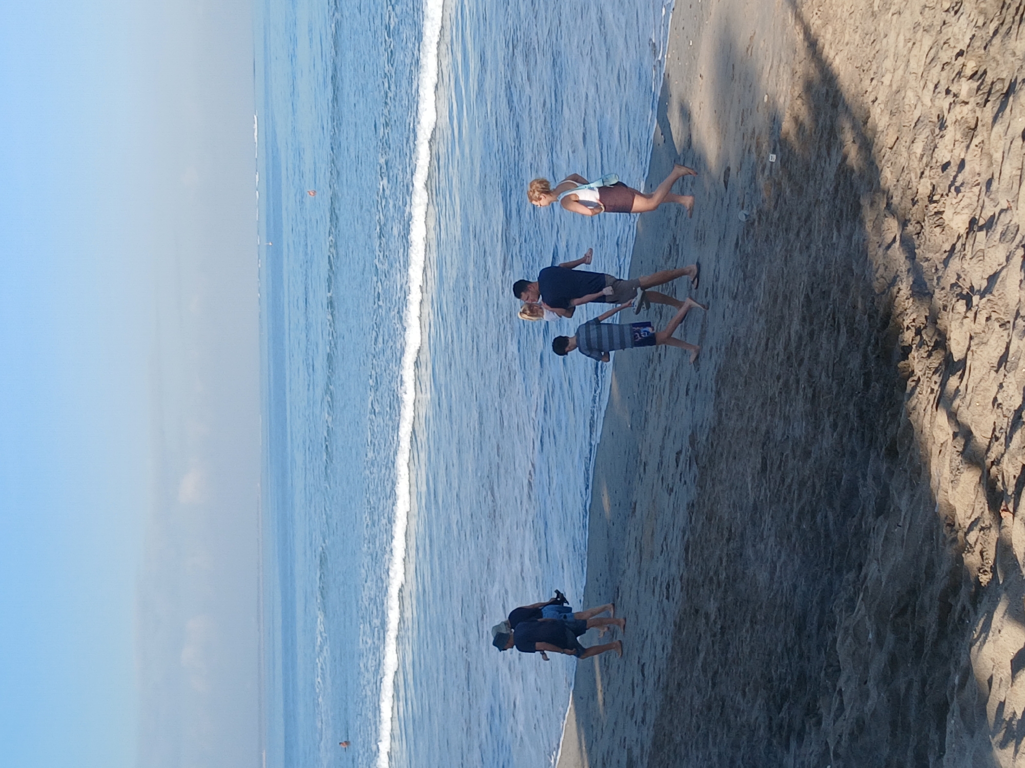 A medium shot captures a group of five individuals walking on a sandy beach adjacent to the ocean in Kuta, Indonesia. The ocean, with small waves, stretches to the horizon, meeting a hazy sky. The foreground consists of sand, casting long shadows from an unseen object to the right.
From left to right:
The first person is a male, wearing a dark t-shirt and dark shorts, with a hat. He is walking away from the camera, towards the water.
To his right is another male, wearing a dark t-shirt and blue shorts. He is walking towards the camera.
Next, a young male, wearing a striped t-shirt and blue swim shorts, walks towards the camera.
Following him is a male carrying a young child. The male is wearing a dark t-shirt and khaki shorts. The child, appearing to be a girl, is wearing a white outfit.
The last person in the group is a female, wearing a white tank top and a dark skirt, walking towards the camera.
The overall scene depicts a relaxed atmosphere on a beach.