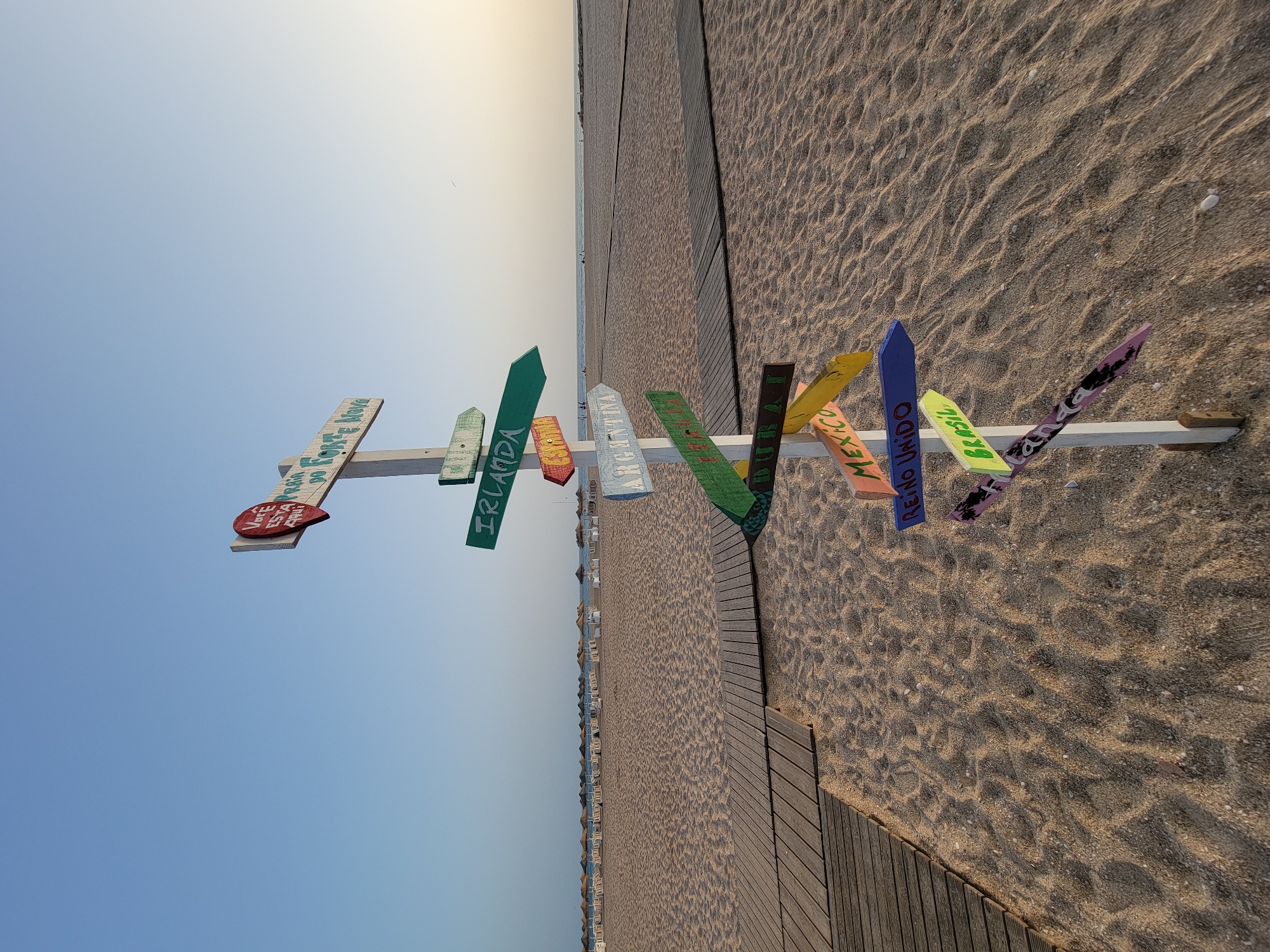 The image shows a directional signpost on a sandy beach in Quarteira, Portugal, with the ocean and sky in the background. The signpost, made of a white wooden pole, has multiple wooden arrows painted with names of various locations. From top to bottom, the visible destinations include: "VOÇÊ ESTÁ EM FORTE NOVA", "IRLANDA", "ESPANA", "ARGENTINA", "REINO UNIDO", "BRASIL", and "CANADA". The beach is lined with rows of beach umbrellas and loungers, suggesting a popular tourist destination. Wooden walkways are laid across the sand. The sky is clear and blue, with the sun setting on the horizon. There are no people clearly visible in the immediate foreground, but a few individuals can be seen in the distance on the beach and near the water. The scene is one of a serene beach environment with a playful, travel-themed artistic installation.