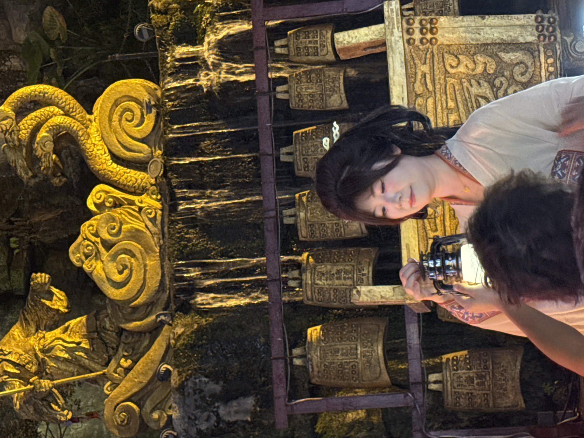 In Yuzhong District, China, a young East Asian woman stands holding a lantern. She is dressed in a traditional light-colored robe with embroidered accents. Behind her, a row of ancient-looking bells hangs from a dark rack. Above the bells, a golden statue depicts a figure holding a staff and a coiled dragon-like creature with multiple heads, situated above a cascading waterfall. The scene appears to be outdoors, possibly a temple or historical attraction, illuminated by artificial light that casts a golden hue.