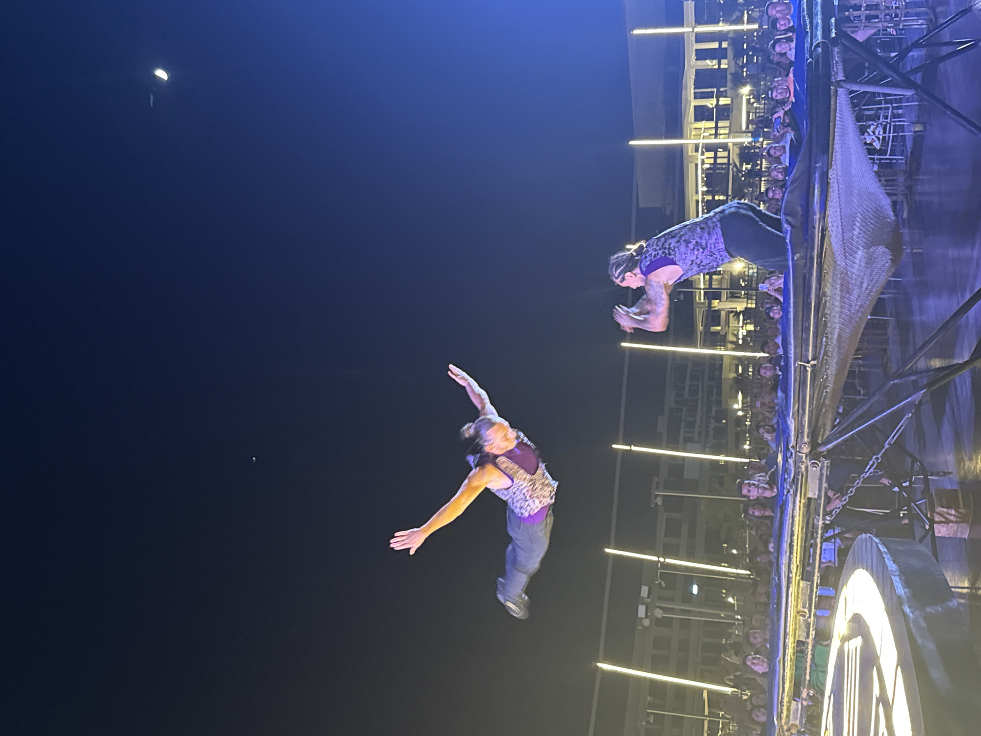 Two acrobats are performing a trampoline act at night in Sharm El Sheikh, Egypt. One acrobat is in mid-air, suspended horizontally with arms outstretched, appearing to be in a diving pose. The other acrobat is on the trampoline, in the process of jumping. Both performers are wearing similar purple and black patterned vests. The trampoline is set up outdoors, with a crowd of spectators watching from behind a barrier. The background features illuminated buildings and light poles, suggesting an urban setting. The sky is dark, with a crescent moon visible.