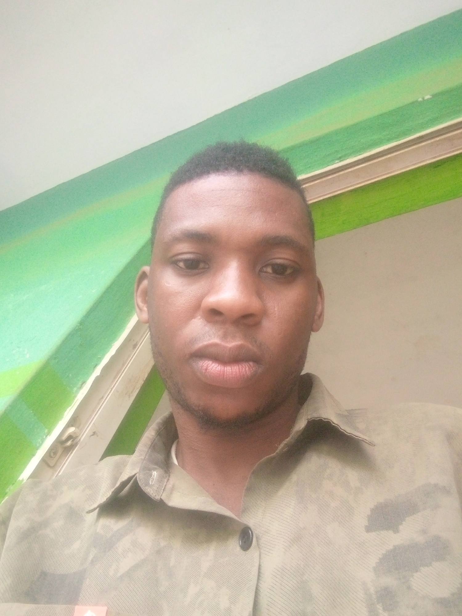 The image is a medium close-up shot of a man with short, dark hair, brown eyes, and dark skin. He is wearing a button-down shirt with a camouflage or textured pattern. The background is a wall painted with a horizontal stripe of green and white, with a trim piece that appears to separate them. The setting seems to be indoors. No specific location or city is identifiable from this image.