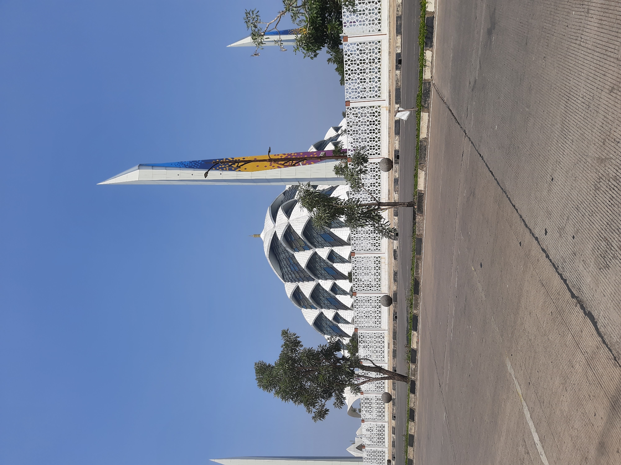 The image captures a facade of the Grand Mosque of Bandung, a prominent religious building in Bandung City, Indonesia. The mosque features a striking white exterior with a distinctive roof structure composed of overlapping, curved panels. Two tall, slender minarets flank the main building, one of which is decorated with colorful geometric patterns and imagery resembling trees. A decorative white fence, featuring intricate latticework, separates the mosque grounds from the street in the foreground. Several small trees are planted along the fence line. The foreground shows a paved road with clear lane markings and a concrete barrier. The sky is a clear, bright blue, suggesting a sunny day. There are no people visible in the image.