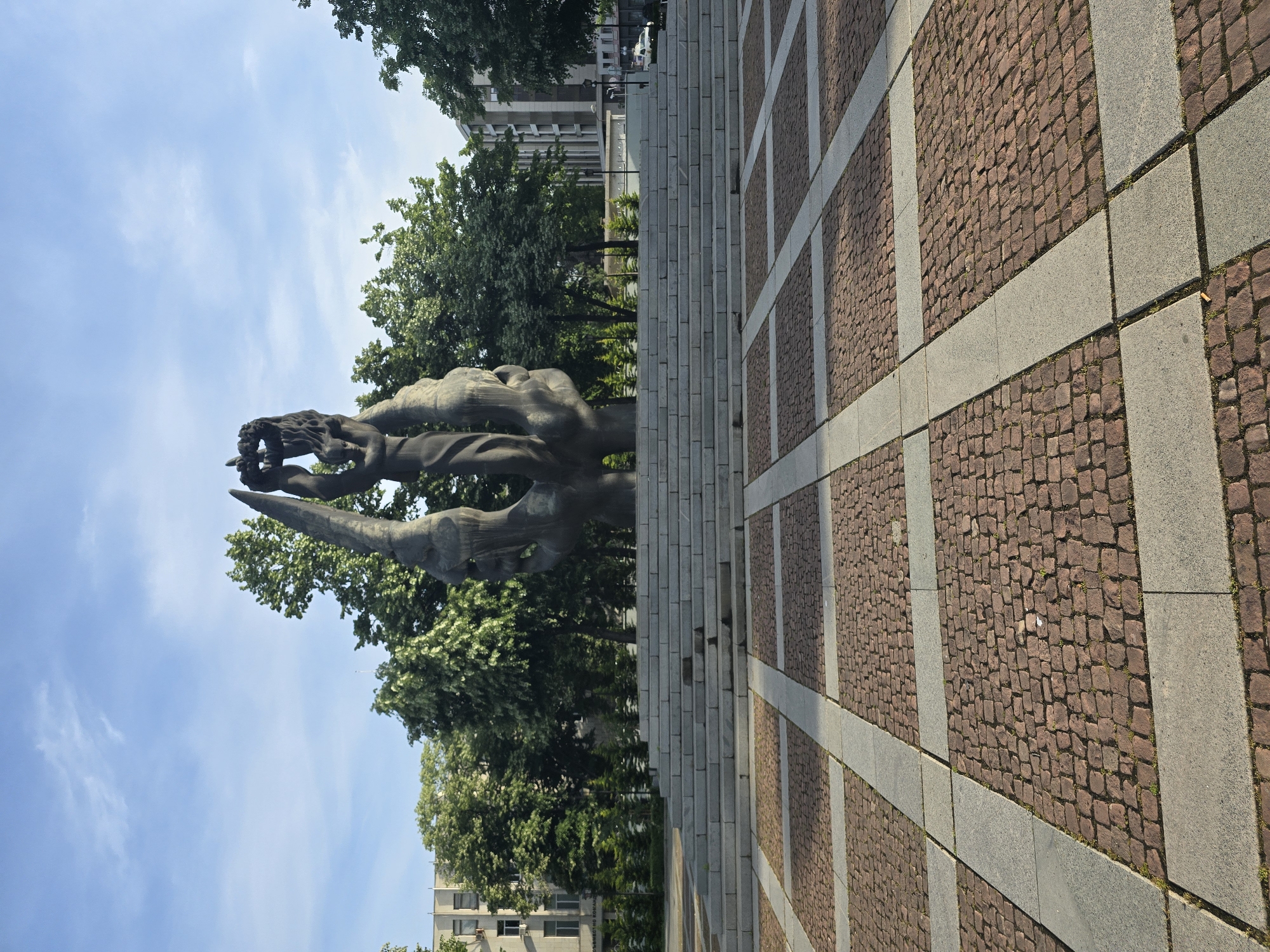 The image shows a vertically oriented view of an outdoor setting. In the foreground, there is a stone-paved area with a grid pattern formed by gray rectangular stones interspersed with sections of reddish-brown cobblestones. Further into the scene, a set of wide stone steps leads up to a large, abstract sculpture made of dark metal or stone. The sculpture appears to depict figures or forms in a stylized manner, reaching towards the sky. Behind the sculpture and to the sides, there are green trees, with some buildings visible in the background. The sky is blue with some wispy clouds. The overall scene appears to be an urban public space with art, architecture, and greenery. The monument is called the Victims of Fascism Monument, located in Skopje, North Macedonia.
