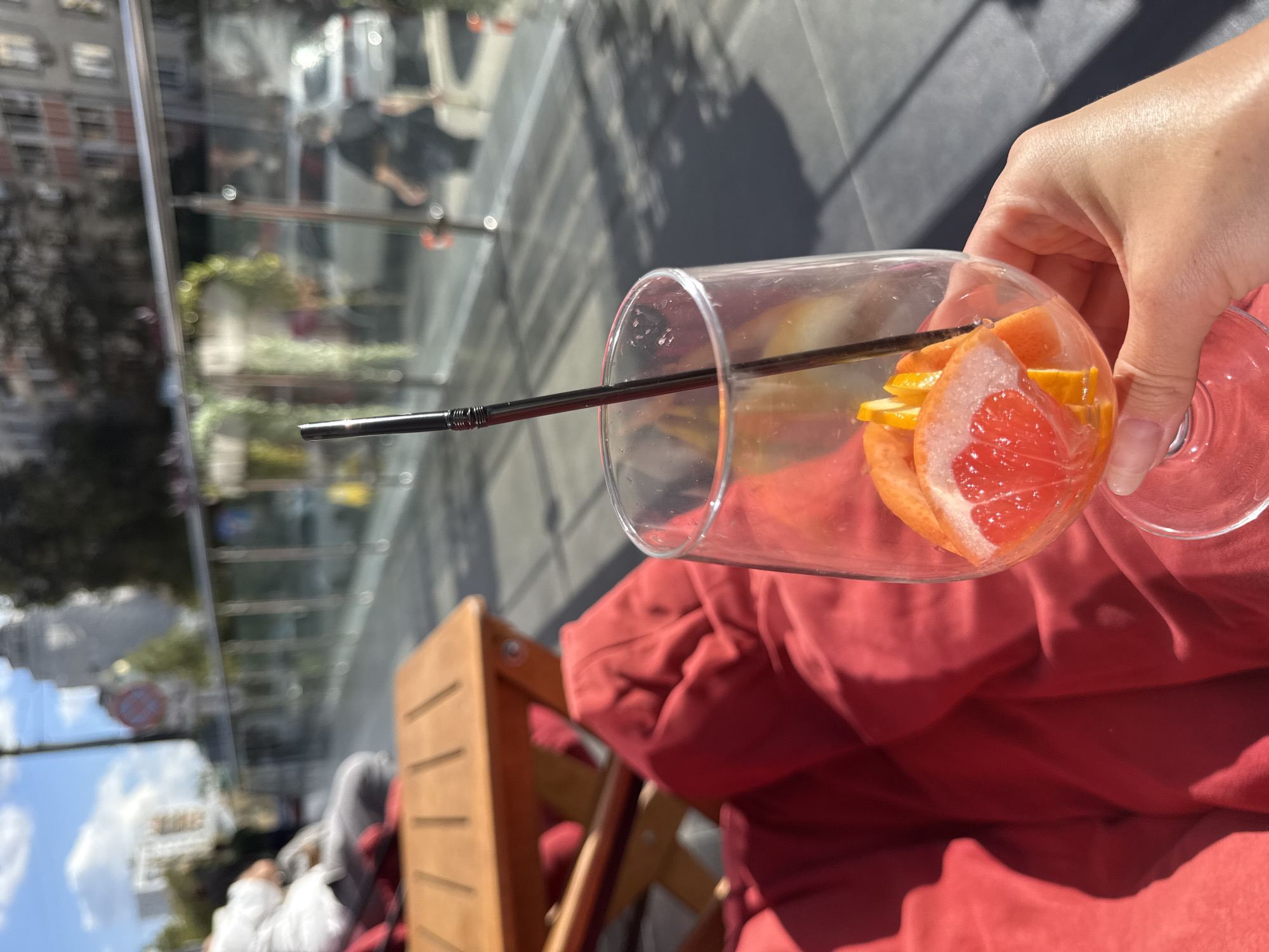 The image depicts a close-up view of a person's hand holding a wine glass filled with citrus fruit slices and a black straw. The glass is positioned in the foreground, with the fruit slices, including grapefruit and orange, clearly visible. Behind the glass, a red cushioned seating area is visible, with a wooden table in the background. The scene appears to be outdoors, with dappled sunlight casting shadows on the ground. In the blurred background, a street or walkway is visible, with people and vehicles in motion, suggesting an urban environment. The architecture in the distance suggests buildings, and a clear sky with clouds indicates daytime. The overall impression is of a relaxed outdoor setting, possibly a cafe or restaurant.