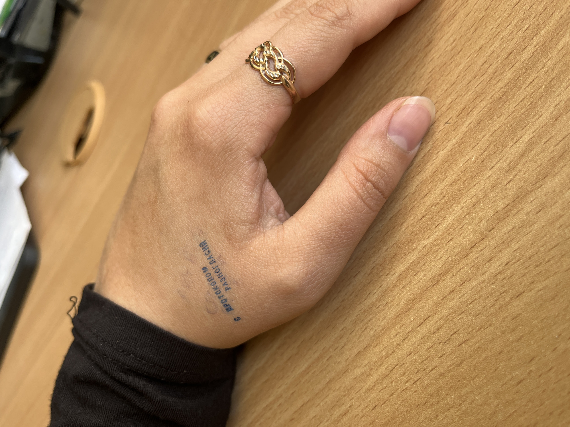 A close-up shot features a person's left hand resting on a light-colored wooden surface, likely a desk or table. The hand is positioned palm down, showcasing the back of the hand.
On the ring finger, a distinctive gold-colored ring is visible, characterized by an intricate, open-work design resembling interwoven loops or a Celtic knot. The fingernails are natural and unpolished. The wrist is adorned with a black long-sleeved garment, possibly a shirt or sweater, partially visible at the bottom left.
A prominent detail is a blue ink stamp on the back of the hand, positioned near the wrist. The text is in Cyrillic script, oriented vertically, and reads "С ПРОТОКОЛОМ РАЗНОГЛАСИЙ". This translates to "WITH A DISAGREEMENT PROTOCOL" or "WITH A PROTOCOL OF DISAGREEMENT."
The wooden surface exhibits a fine grain pattern throughout the image. In the blurred background to the upper left, glimpses of other objects, such as white paper or fabric, are faintly discernible. The setting appears to be indoors, possibly an office or study area, given the wooden surface and the nature of the hand stamp. No specific activity is taking place other than the hand resting.