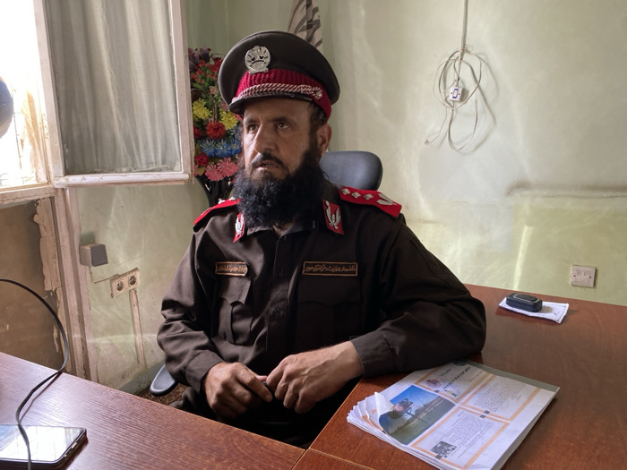 The image depicts a middle-aged male with a dark, full beard and mustache, seated behind a reddish-brown wooden desk in an office setting within the city of Unknown, Unknown. He wears a dark brown military-style uniform, consisting of a peaked cap with a red band and silver insignia, and a shirt with shoulder boards featuring red piping and silver insignia, along with breast patches displaying white Arabic script. The man's gaze is directed off-camera to the left. On the desk, a smartphone with a black cable lies on the left. A stack of printed documents, including visible text and a landscape image, is centered in the foreground. A small, dark rectangular electronic device rests on a piece of folded white paper to the right. The background features light green-beige walls with visible stains and wear. A white-framed window with a translucent curtain is on the left, below which exposed electrical outlets and wires are mounted. An artificial floral arrangement in a dark vase is positioned behind the man's head, and a bundle of white electrical wires with a small white box is affixed to the wall above his right shoulder. Another electrical outlet is visible on the right wall. The man is seated in a dark office chair.