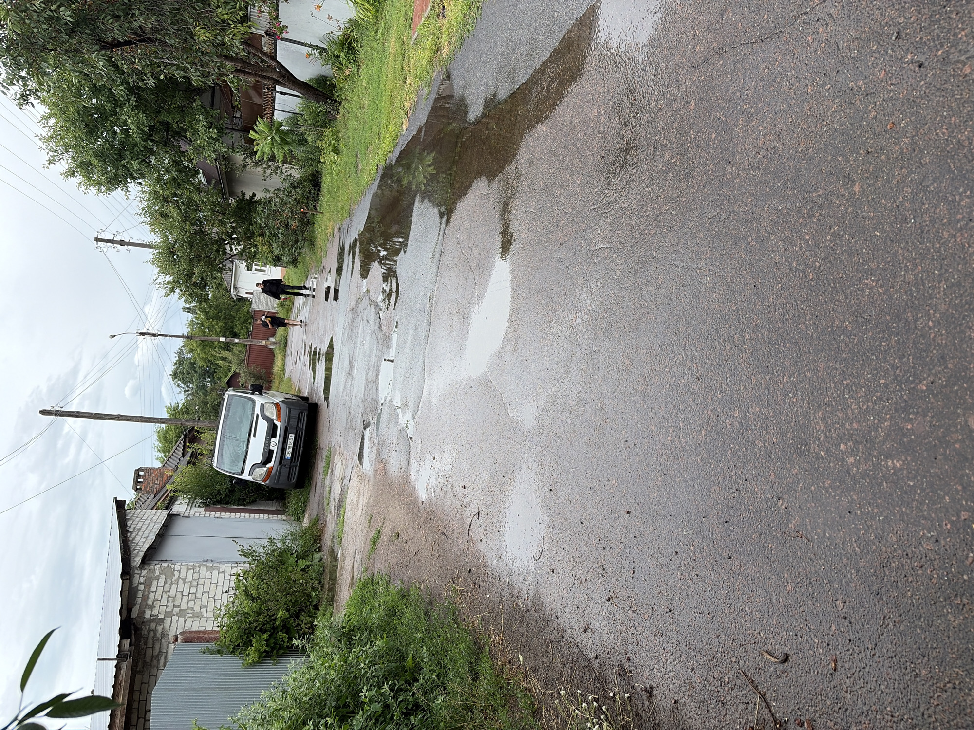 A residential street in Chernihiv, Ukraine, is depicted under an overcast sky. The asphalt road is wet and shows numerous puddles, indicating recent rain. A white van is parked on the left side of the road, near a brick building with a metal garage door and a corrugated metal fence. Green foliage and trees line the sides of the street. Two individuals are walking away from the camera on the right side of the road. The scene suggests a typical day in a Ukrainian town, possibly after a rain shower.