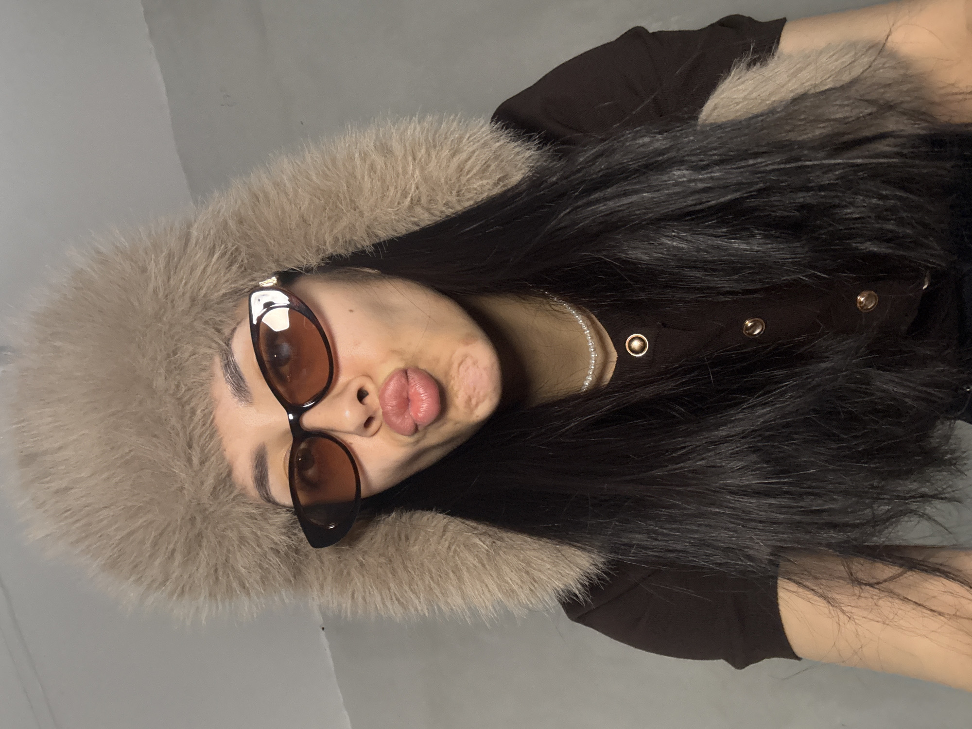 A young woman is captured in a medium close-up, tilted portrait against a plain, light grey background. She has long, straight dark hair that flows around her. Her face is framed by a voluminous, light brown or beige faux fur-trimmed hood or collar. She wears dark-lensed, cat-eye style sunglasses and has her lips playfully puckered into a "kissing" or "duck face" expression. Her attire includes a dark brown or black top, possibly a jacket, detailed with prominent round, gold-colored buttons or snaps down the front. A delicate necklace of white pearls or beads is visible around her neck. The lighting is soft and even, suggesting an indoor setting. The overall mood conveyed is playful and confident. No text is visible in the image. The location context for this media file is Votkinsk, Russia.