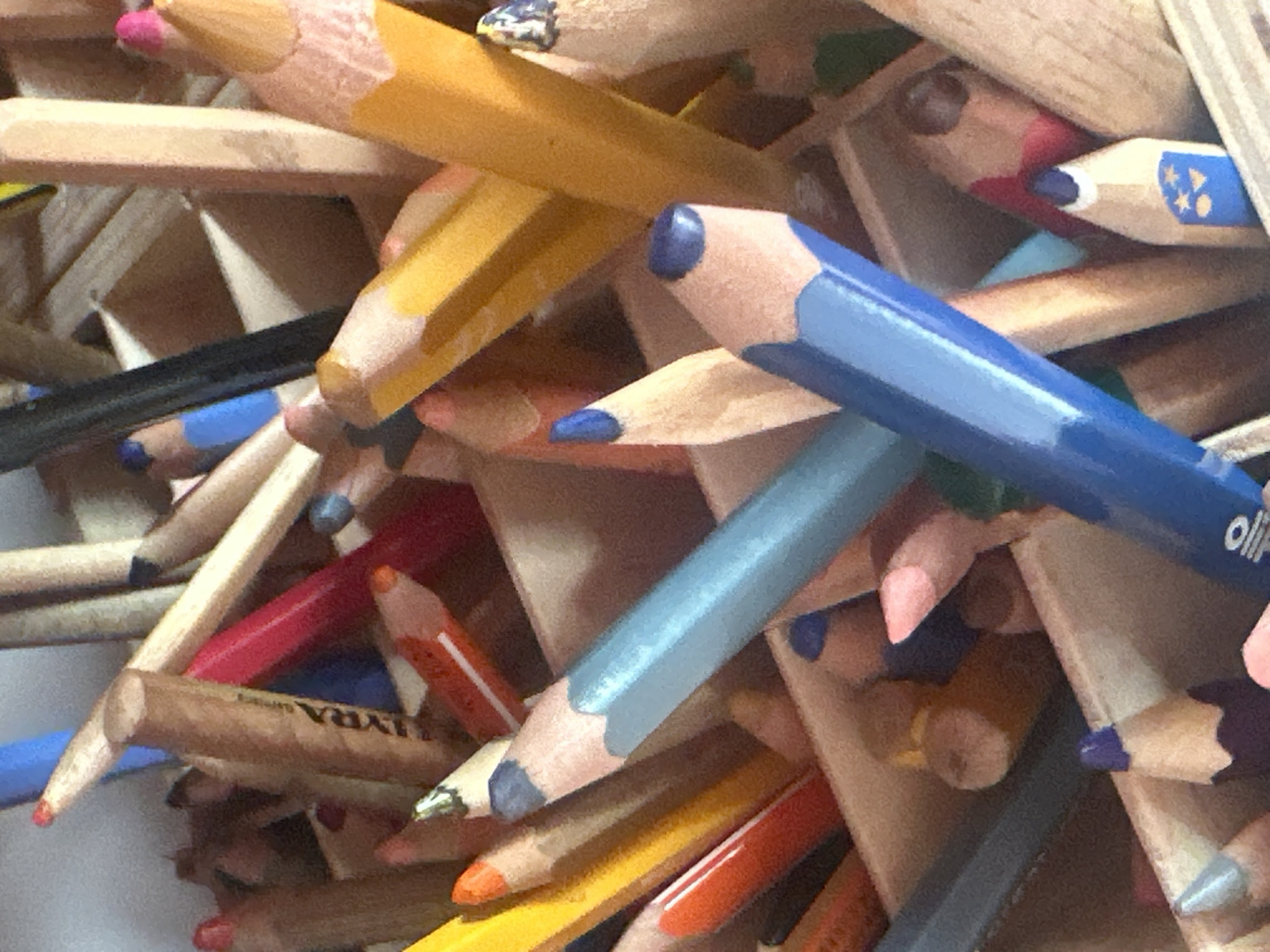 A close-up, eye-level shot shows a wooden organizer filled with sharpened colored pencils. The pencils are tightly packed, with their points facing upwards and their shafts angled in various directions. Colors visible include yellow, blue, red, orange, and natural wood. Some pencils have visible brand markings, such as "LYRA". The background is blurred but suggests a soft, diffused light source. The setting is indoors, and the overall impression is one of an art or craft supply storage area. There are no people or discernible actions taking place.