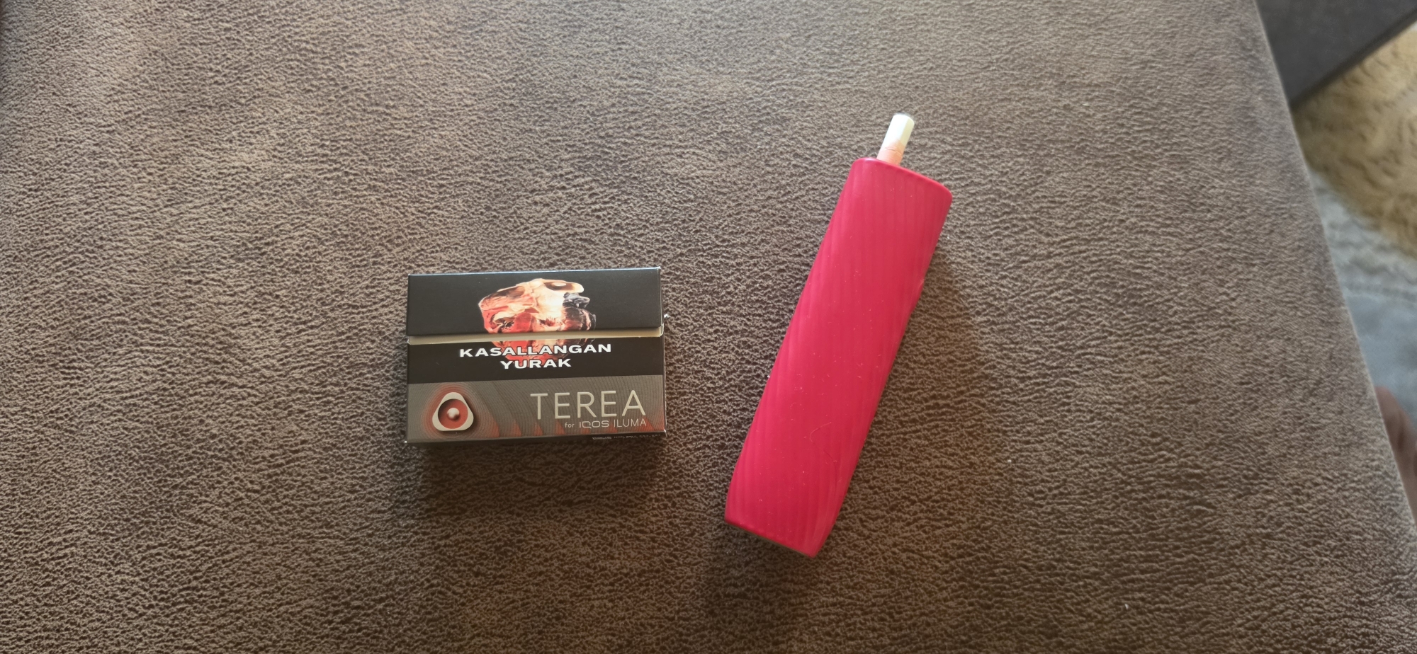 The image shows a close-up, overhead view of two objects on a textured brown surface. On the left is a black and silver pack of "TEREA" tobacco sticks for "IQOS ILUMA," with the words "KASALLANGAN YURAK" printed above the brand name. A red circular graphic with a white center is visible on the lower left of the pack. To the right of the tobacco pack is a bright pink, ribbed cylindrical device, possibly a vape or e-cigarette, with a white tip. The background is a dark brown, textured fabric. The location is not visible in the image.