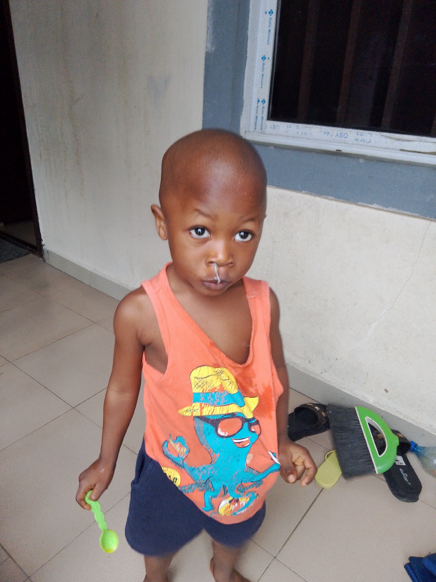 A young boy with a bald head stands in front of a light-colored wall and tiled floor, facing the camera. He is wearing an orange sleeveless shirt with a graphic of a blue octopus wearing a hat and sunglasses, and dark blue shorts. A small amount of mucus is visible on his upper lip. In his left hand, he holds a bright green plastic spoon. To his right, on the floor, are a pair of black sandals with a Louis Vuitton logo, a green object that appears to be a dustpan, and a clear plastic bottle. The background includes a white-framed window with dark vertical blinds. The overall environment appears to be an outdoor or semi-outdoor residential area in Yenegoa, Nigeria.