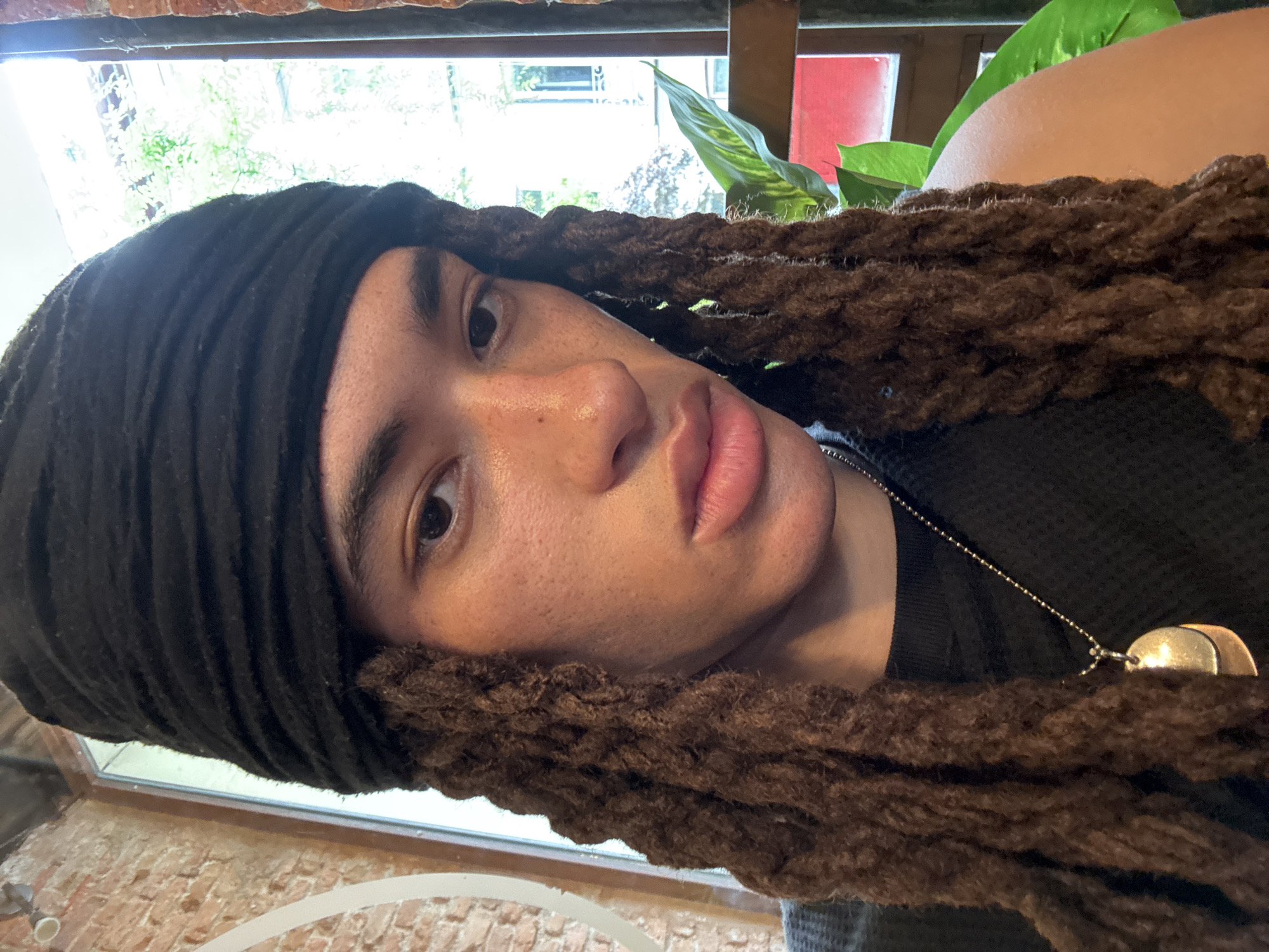 A close-up portrait shows a person with a medium skin tone and dark brown dreadlocks, pulled back under a black headwrap or durag. The subject is oriented sideways within the frame, with their face angled slightly upwards and their gaze directed straight towards the camera. They have dark eyes, full lips, and clear skin with visible freckles. A golden pendant hangs from a thin chain necklace around their neck, partially visible over a black shirt.
The background is softly lit by natural daylight coming through a window, visible behind and above the subject's head. Outside the window, green foliage from plants or trees can be seen, along with glimpses of a building that includes a red-colored section. Inside, elements of brickwork are visible in the lower portion of the frame, suggesting an interior setting. Another green plant is partially visible to the right of the person's head. The overall impression is a casual, intimate portrait taken indoors during the day. There is no visible text in the image. The scene is set in Madrid, Spain.