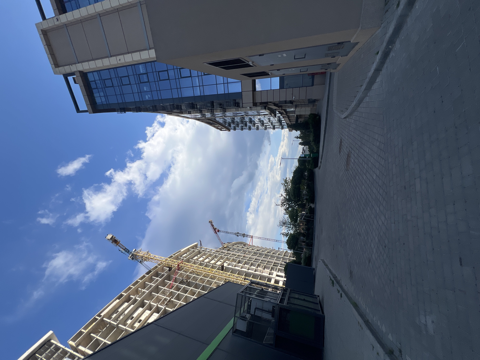 The image shows an exterior view of a construction site and a modern building on a sunny day with scattered clouds in a city. In the foreground, a paved walkway with interlocking grey bricks leads into the scene. To the left, a dark grey building with large glass structures is partially visible, with a green stripe running along its side. Further back and to the left, a large building under construction is seen, featuring a concrete frame and multiple floors. Yellow and red construction cranes are prominently positioned near this building. To the right of the walkway, a modern building with a facade of blue glass windows and beige panels stands tall. The background reveals trees and a clear sky with white clouds. The setting appears to be in the city of Fontanka, Ukraine. No people or animals are visible in the frame.
