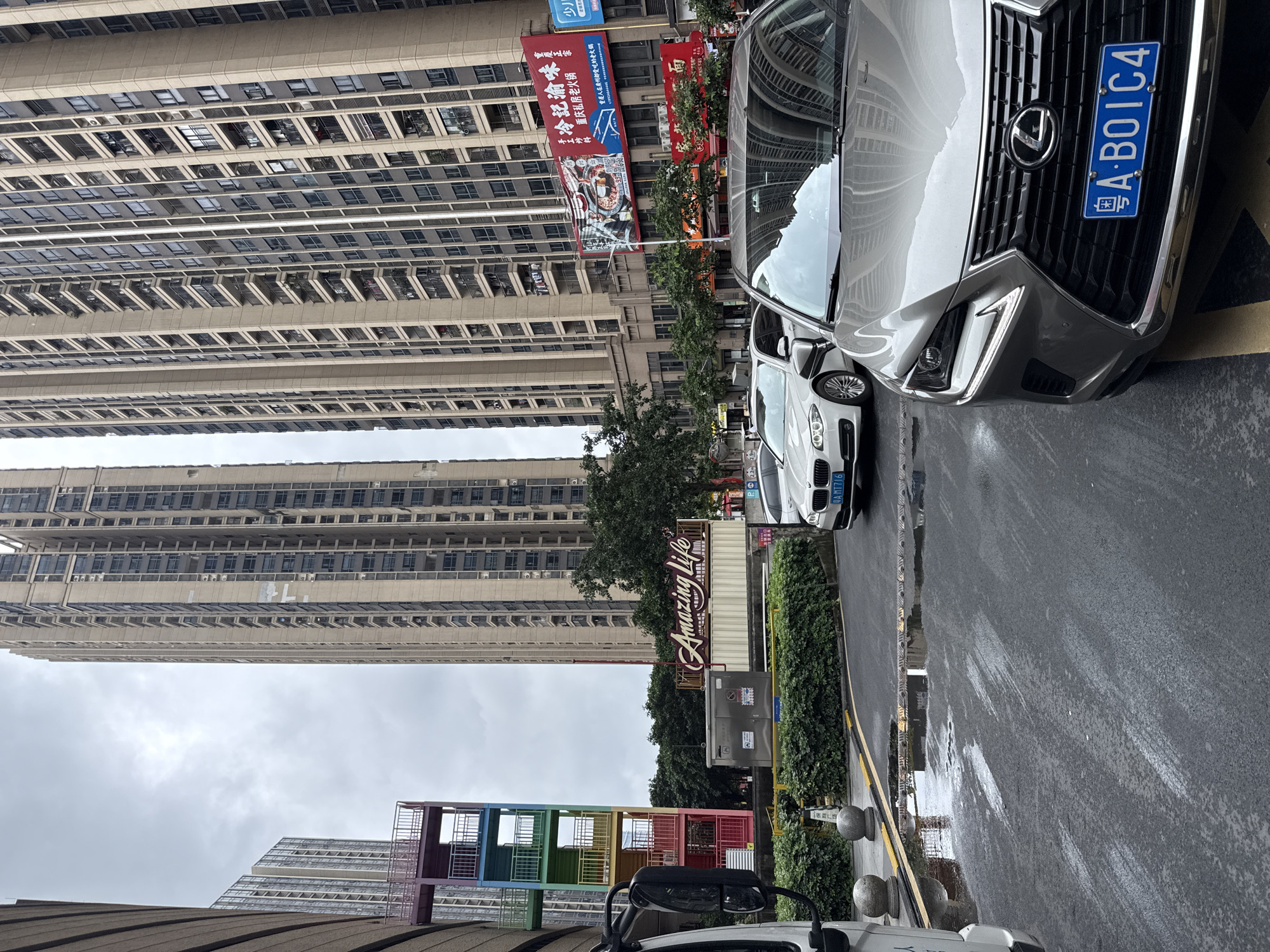The image depicts a street scene in Haizhu District, China, on a cloudy day. In the foreground, a silver Lexus NX 300h is parked on a wet asphalt road, with its license plate reading "A B01C4". To its left, a white BMW sedan is also parked. A white truck is partially visible on the far left. The road has some puddles and white paint markings.
Behind the vehicles, a lush green hedge lines the area. Above the hedge, a large industrial-style shipping container is adorned with a sign that reads "Amazing Life" in red stylized lettering. Adjacent to the container is a grey electrical or utility box.
Further back, colorful external staircases with railings, painted in red, orange, yellow, green, blue, and purple, are attached to a building on the left.
Dominating the background are two exceptionally tall, modern residential skyscrapers with many windows and balconies. Their beige-toned facades suggest a contemporary architectural style. Several other buildings, including one with a large red billboard featuring Chinese characters and images of food, are also visible. The sky is overcast with diffused light, indicating a lack of direct sunlight. No people are prominently featured in the scene.