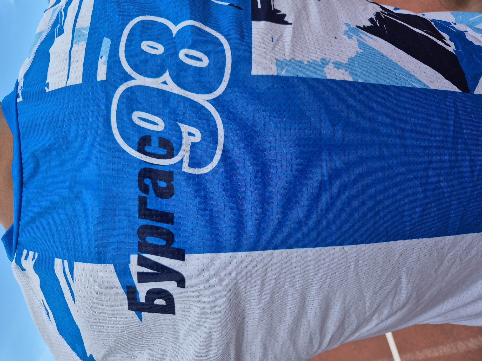 A close-up, low-angle shot shows the back of a person wearing a white and blue athletic shirt with a graphic design. The text "Бургас 98" is prominently displayed in a dark blue font on a blue and white striped section of the shirt. The background is out of focus but appears to be an outdoor setting with a hint of blue sky at the top and an orange-colored surface, possibly a track, at the bottom. The fabric of the shirt has a visible texture with small perforations.