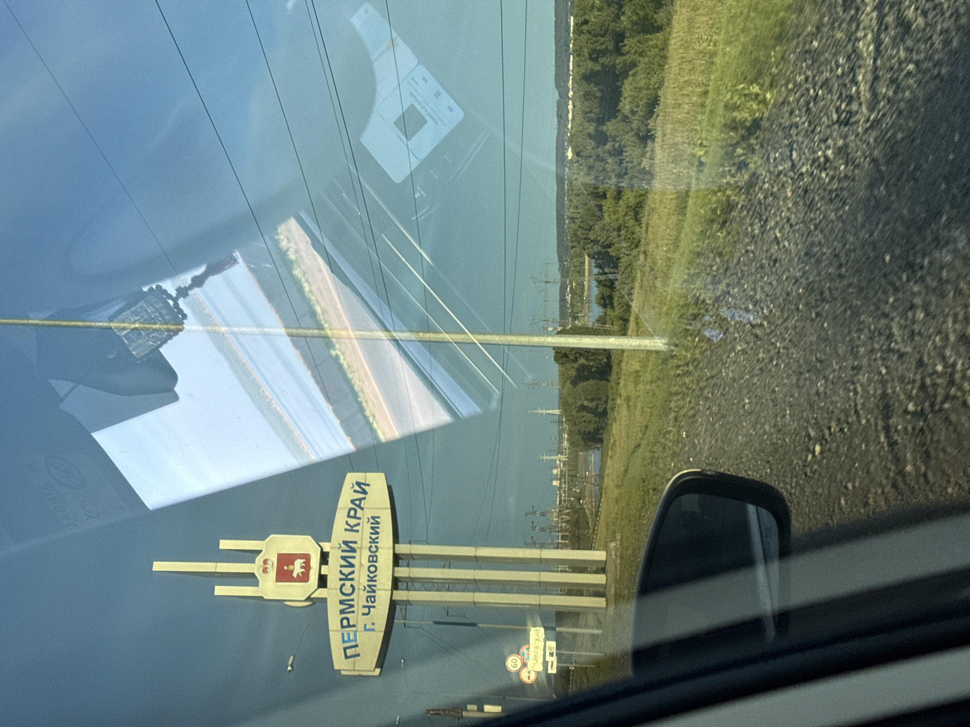 The image captures a roadside scene from within a vehicle, primarily featuring a large, light-colored sign marking an entry point into a region and city. The sign prominently displays "ПЕРМСКИЙ КРАЙ" (Perm Krai) and "Г. ЧАЙКОВСКИЙ" (Chaikovsky city) in Cyrillic, accompanied by a coat of arms depicting a bear, characteristic of the Perm region. Below this main sign, a smaller road sign indicates a 60 km/h speed limit and includes partially obscured text that appears to reference Chaikovsky again.
In the background, a vast body of water, likely a reservoir or large river, stretches beneath a clear, bright blue sky. It is bordered by a dense line of green trees and distant industrial structures, including power lines and pylons. A gravel or dirt road shoulder is visible to the right.
The foreground is dominated by reflections on the car's window. These reflections include portions of the vehicle's dashboard, a decorative hanging charm, and a clear reflection of a smartphone screen. On the phone screen, media player controls (play/pause, skip forward/backward) are visible, along with the Cyrillic text "добавлено в плейлист" (added to playlist). A side mirror of the car is also partly seen on the far right. The photograph was taken during the daytime, under sunny weather conditions, and depicts a traveler entering or passing through Chaikovsky, Russia.