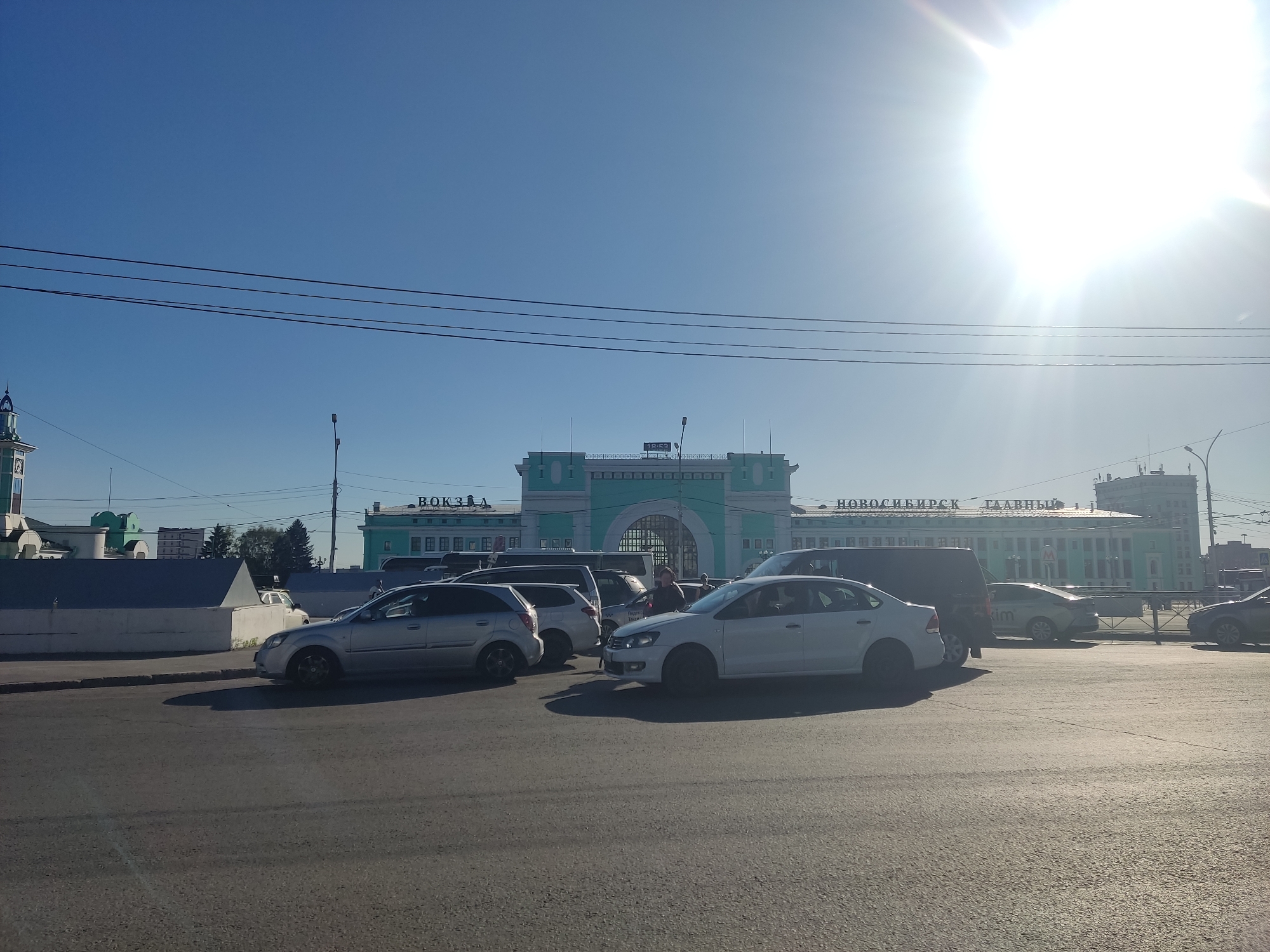 The image captures a daytime scene in front of the Novosibirsk-Glavny railway station in Novosibirsk, Russia. The bright sun is high in the sky, casting long shadows and creating lens flares. Several cars are parked or driving on a paved road in the foreground, including a silver sedan, a white sedan, and a dark SUV. A few people are visible near the vehicles, including one standing beside the white sedan. In the background, the distinctive teal and white architecture of the railway station is prominent, with "НОВОСИБИРСК ГЛАВНЫЙ" (Novosibirsk Main) signage visible. Utility poles and wires are strung across the sky. The overall atmosphere is that of a busy urban transportation hub on a sunny day.