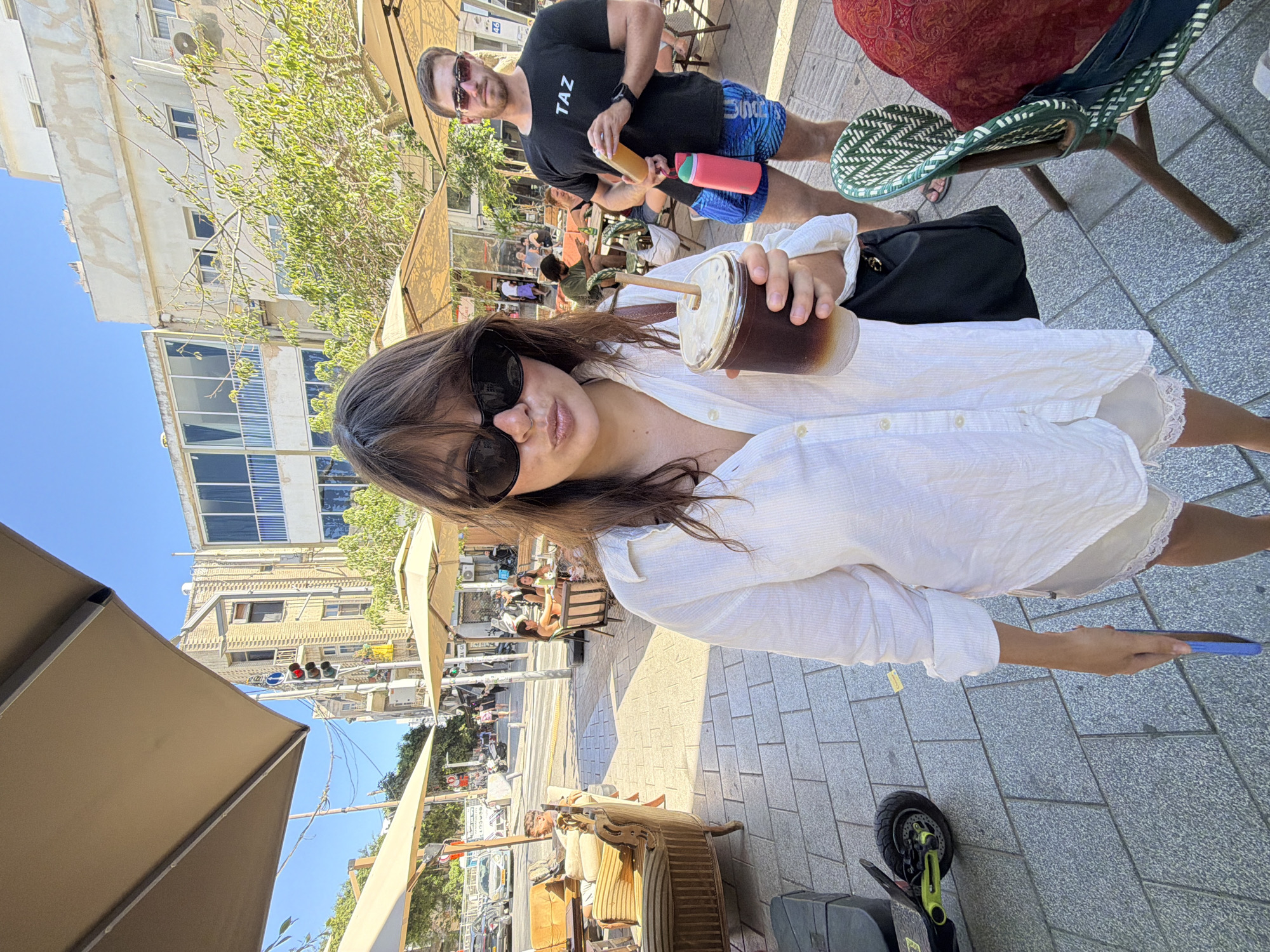 A woman is holding a clear plastic cup with a straw, containing a dark beverage with foam. She is wearing large black sunglasses and a white button-down shirt. The setting appears to be an outdoor cafe or restaurant with paved ground and umbrellas providing shade. In the background, a man wearing a black t-shirt with white text and blue patterned shorts is visible, holding two bottles, one pink and one orange. Other people are seated at tables in the background. The scene suggests a casual outdoor gathering on a sunny day. The text on the man's shirt reads "TAZ". The location cues, such as the architecture and traffic light, are consistent with Tel-Aviv, Israel.