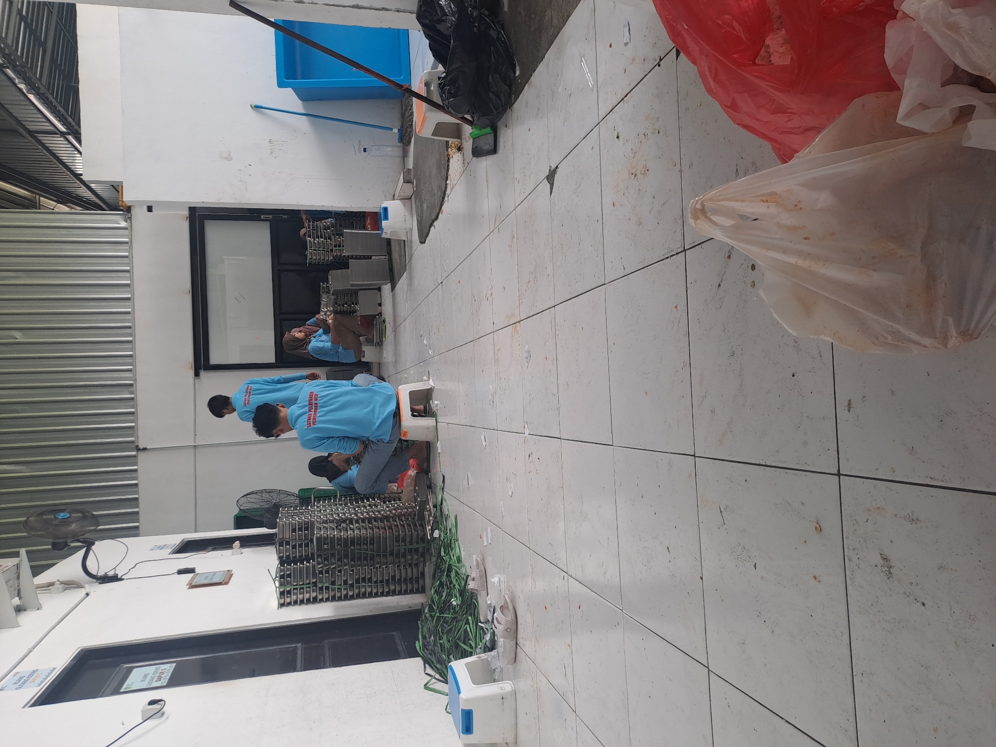 The image depicts an indoor scene, likely a warehouse or workshop, in Mojorejo, Indonesia. Three individuals, appearing to be workers, are present. One individual is standing and facing away from the camera, wearing a light blue long-sleeved shirt with text on the back. Two other individuals are seated on the floor or low stools, also wearing light blue shirts. One seated individual is facing the camera and appears to be working on something on the floor. Another seated individual is partially visible and facing away.
The environment is characterized by a tiled floor that appears somewhat dirty with scattered debris. Stacks of metal trays or containers are visible against a white wall. Various objects are scattered around, including plastic stools, a green bundle of what appears to be rope or cable, a blue plastic bin, a broom, and several black trash bags. The background shows a corrugated metal ceiling and white walls. A large window or glass partition is also visible on the right side of the frame. The overall impression is of a functional but somewhat disorganized workspace.