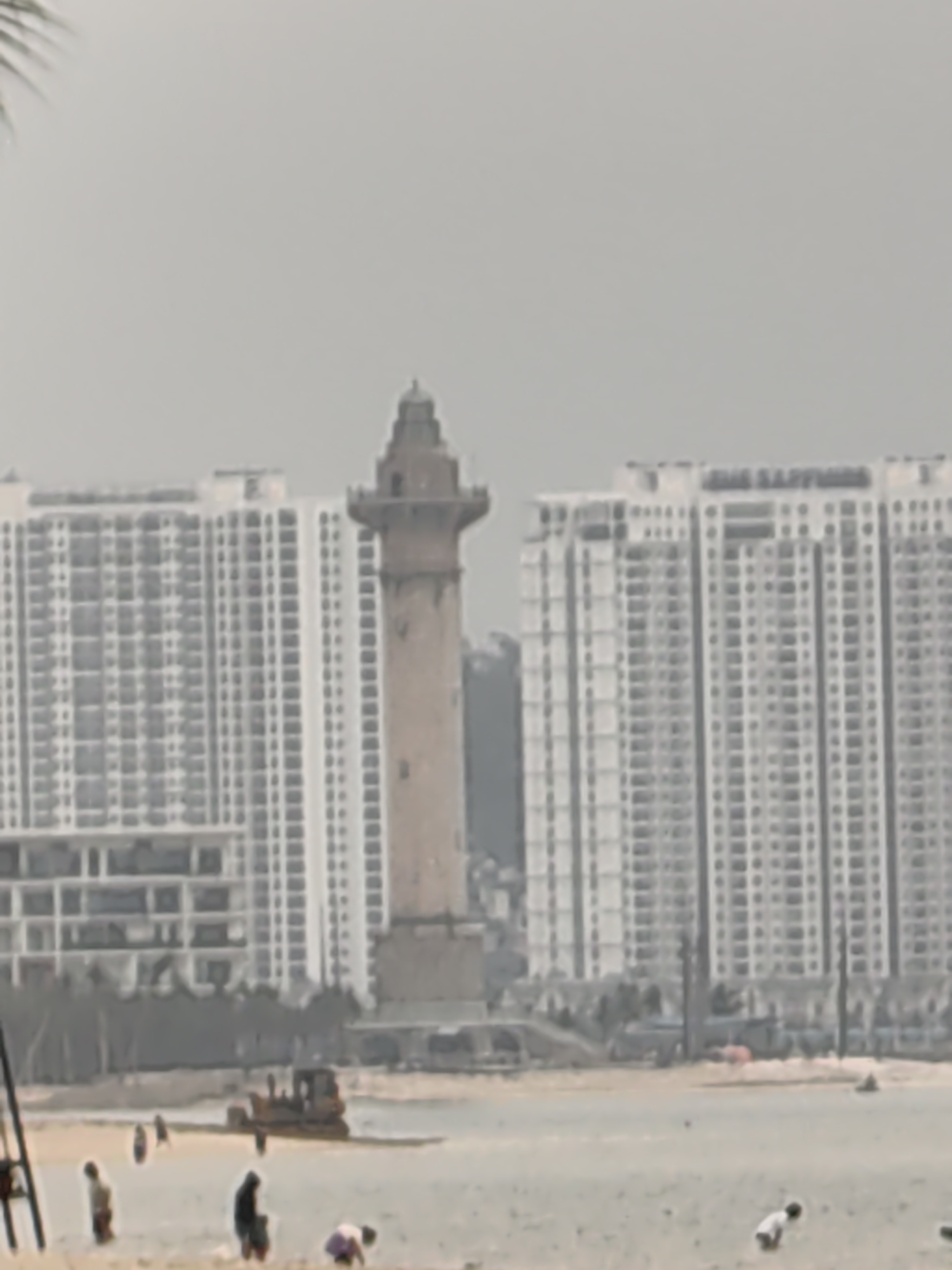 The image depicts a hazy coastal scene in Bai Chay Ward, Vietnam. In the foreground, several individuals are scattered along the beach, some standing, others bending over, and one appearing to be swimming. A construction vehicle, possibly a bulldozer, is positioned on the sand near the shoreline. The background is dominated by high-rise apartment buildings with numerous windows. Between these buildings, a tall, cylindrical tower with a distinctive architectural style and a pointed roof rises, seemingly a historical or landmark structure. The overall atmosphere suggests a blend of tourism, construction, and residential life.