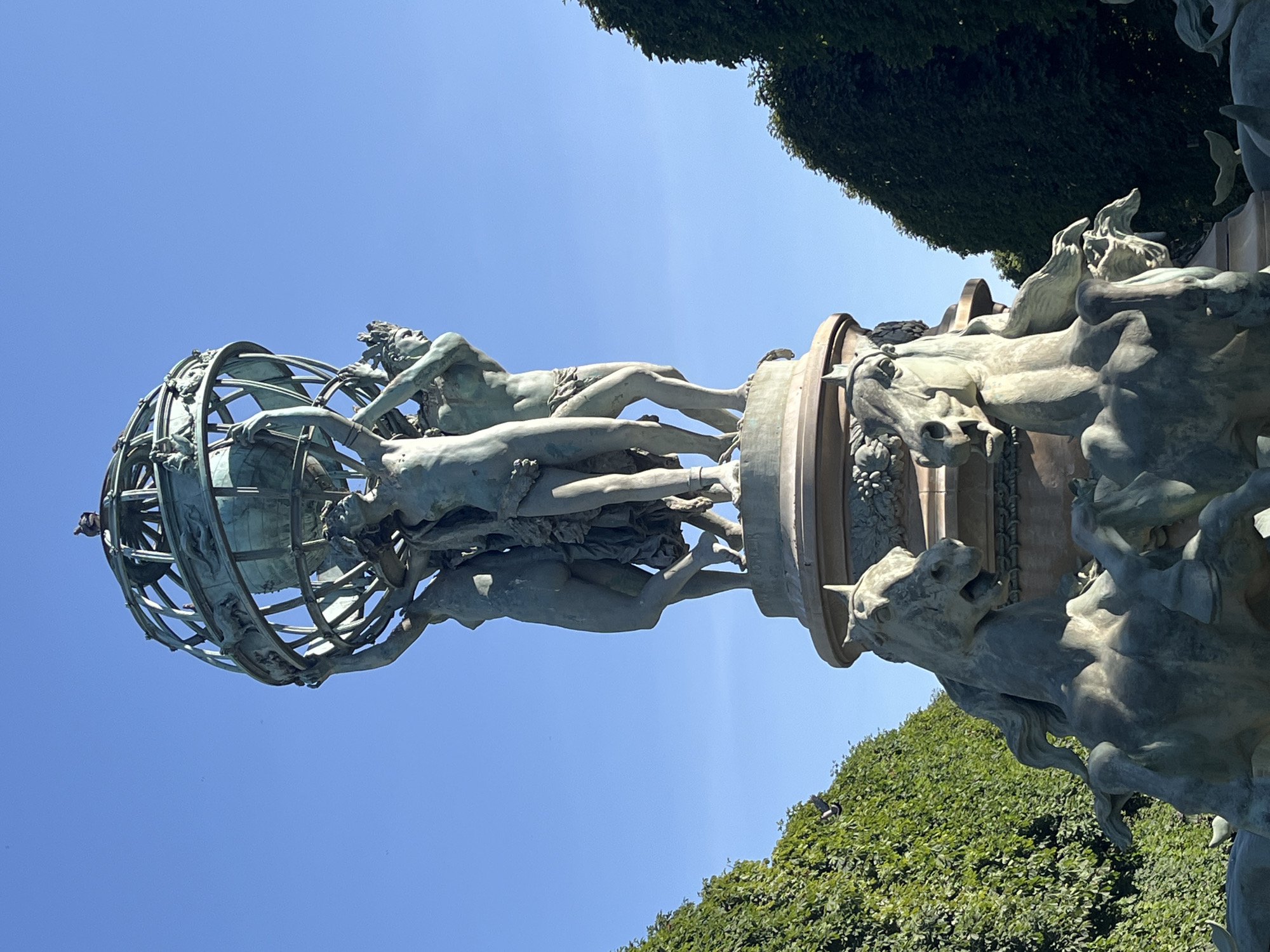 The image shows a bronze sculpture featuring a globe supported by three figures, set against a clear blue sky. The globe is intricate, with a latticed structure adorned with depictions of animals and figures. The three supporting figures are muscular and appear to be lifting the globe collectively. Below this central element, the sculpture transitions to a base adorned with more figures, including what appear to be horses with dramatic expressions, possibly part of a fountain. The sculpture is surrounded by lush, manicured green hedges that frame the scene on either side. The lighting suggests a sunny day, casting shadows and highlighting the details of the bronze work.
This sculpture is the Fontaine de la Concorde, located in the Place de la Concorde in Paris, France. The fountain was designed by Jacques Hittorff and built between 1835 and 1840. It is one of two identical fountains in the square, the other being the Fontaine des Mers.