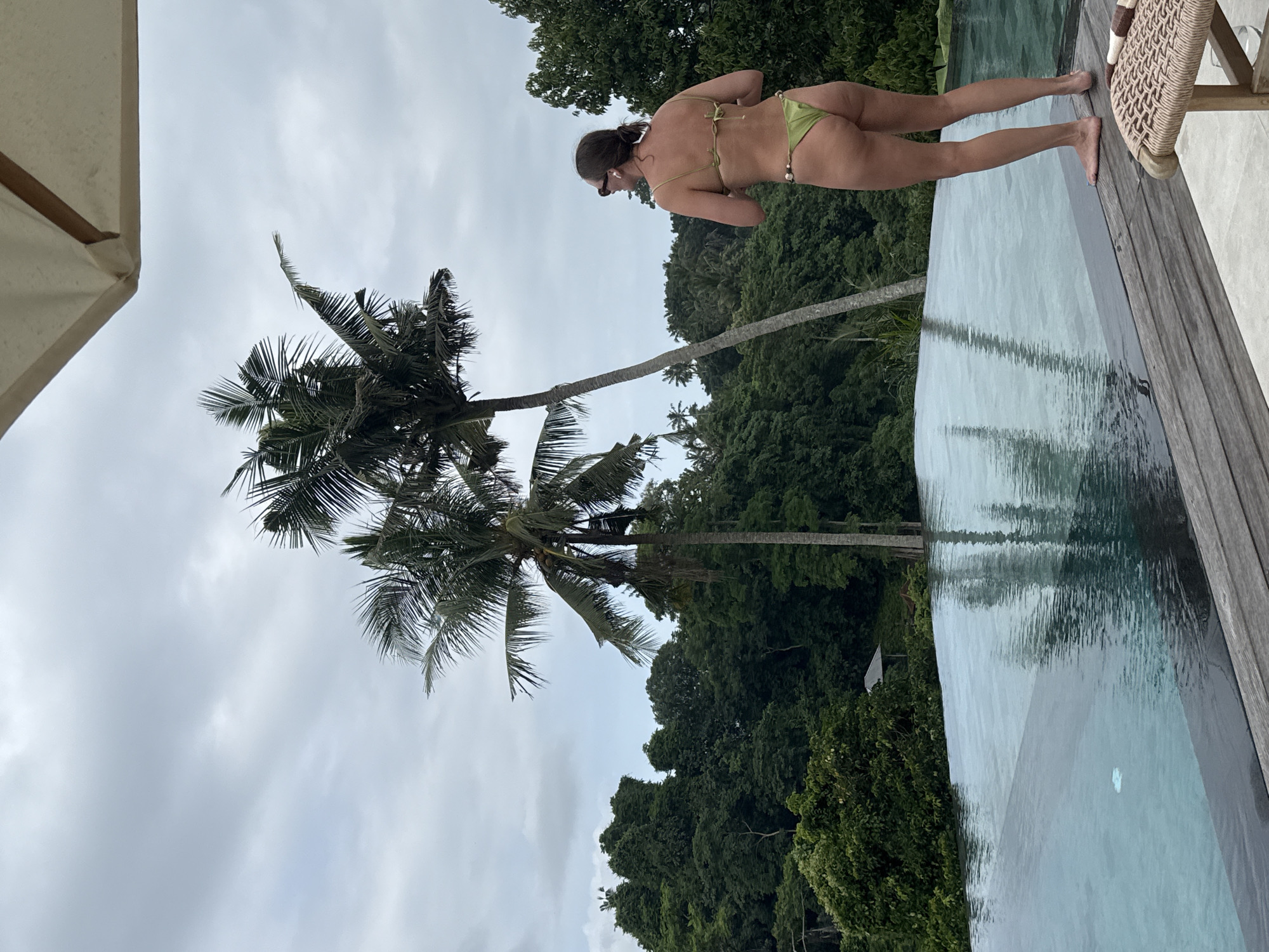 A waist-up view of a woman in a green two-piece swimsuit standing on the edge of an infinity pool. The pool is surrounded by wooden decking. Beyond the pool, there is dense tropical forest. A palm tree is on the left side of the frame, and the sky is overcast. The location is the Ubud District, Indonesia. A beach chair is on the right side of the frame.