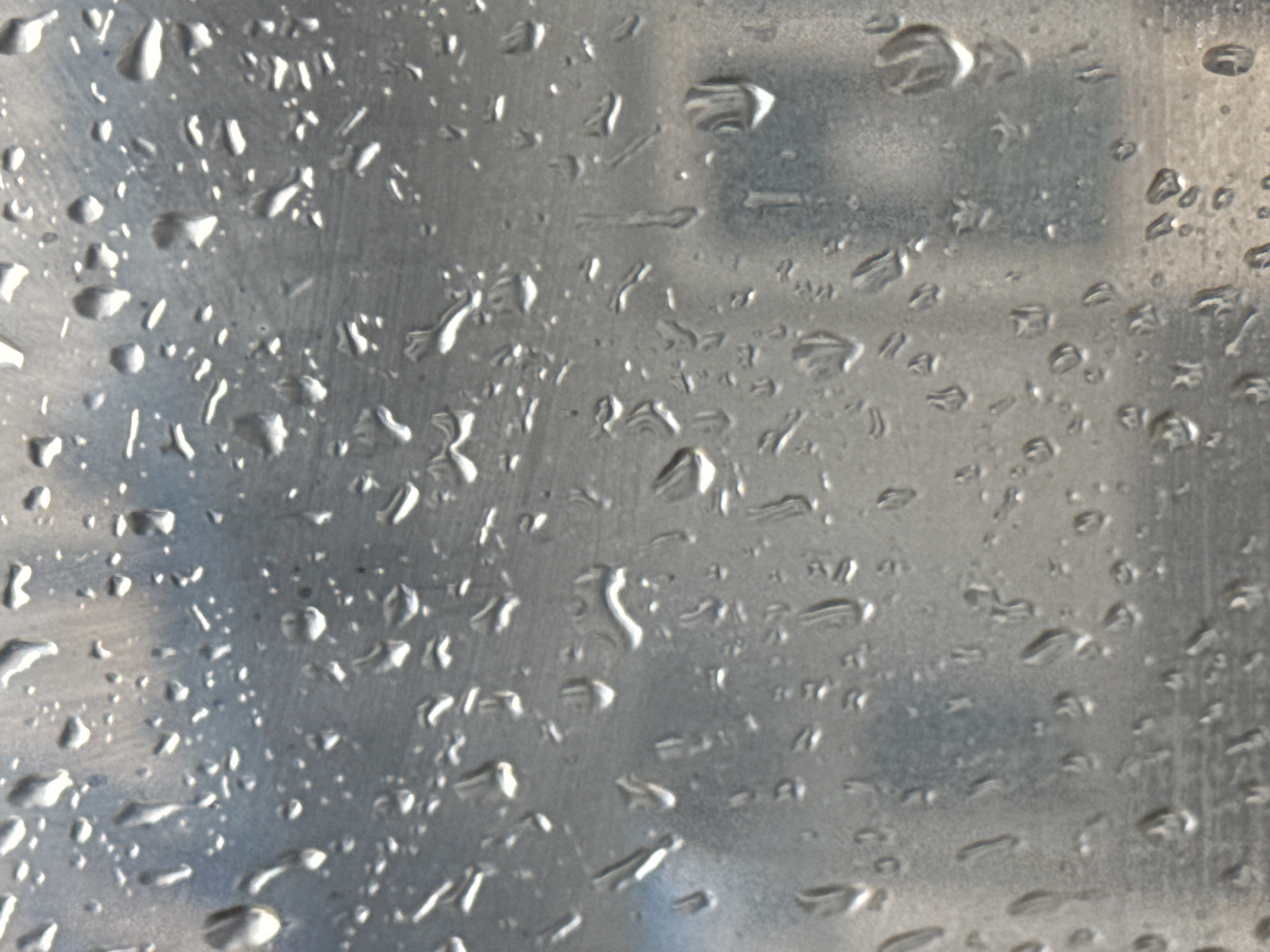 The image shows a close-up view of raindrops on a metallic surface, likely a window or a metal panel. The drops vary in size and shape, with some appearing as small beads and others as larger, more elongated streaks. The metallic surface has a brushed or slightly textured finish, and the reflections within the water droplets suggest an outdoor environment, possibly under a cloudy sky or diffused light. There are no people or other discernible objects in the foreground, and the background is blurred and indistinct, making it impossible to identify specific urban features of Tiraspol, Moldova. The image captures a moment of precipitation and its effect on a man-made surface.