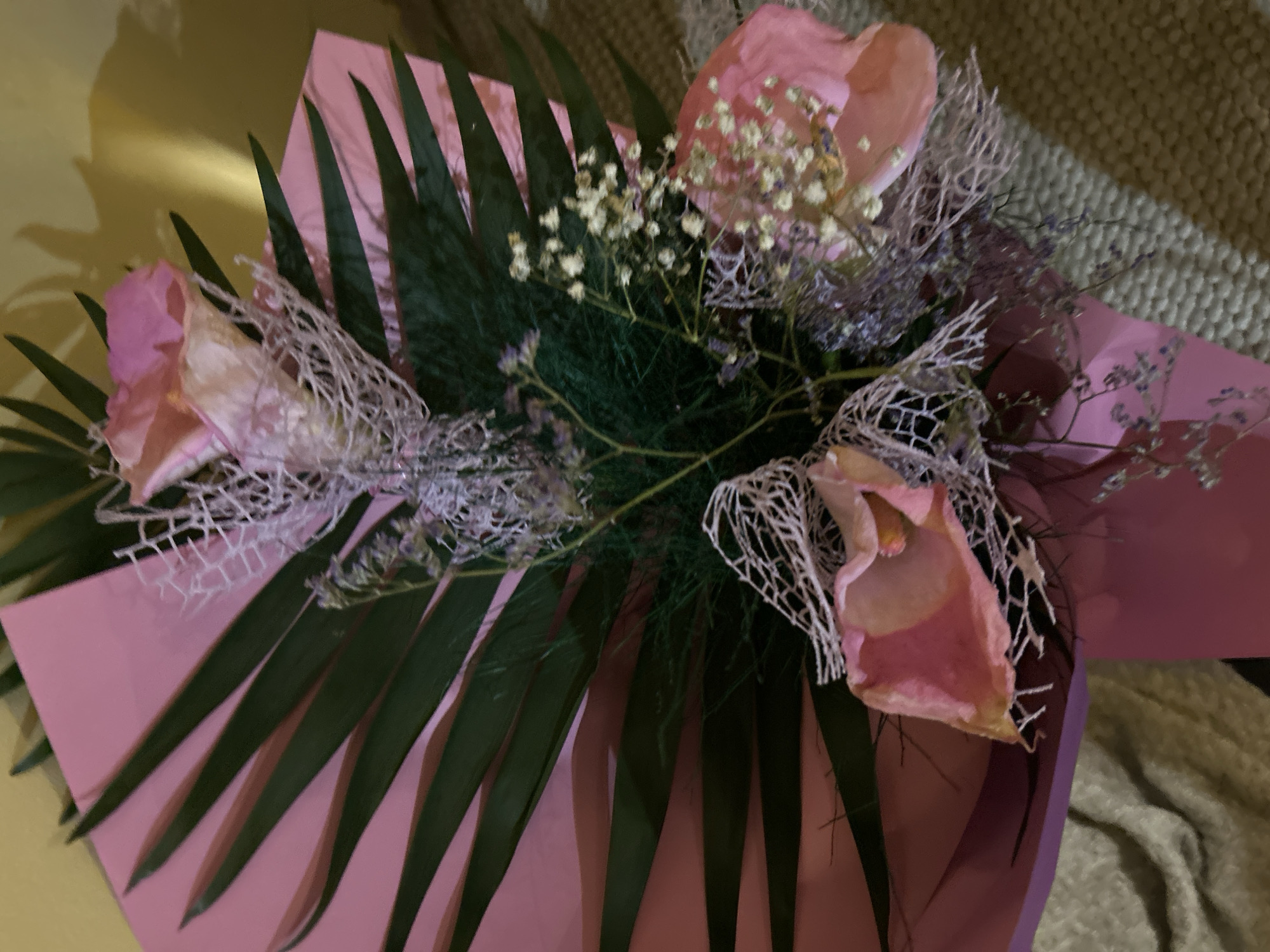 A close-up overhead shot depicts a floral arrangement, likely a bouquet, wrapped in pink and purple paper. Three prominent pink flowers, possibly roses or a similar variety, are visible. These are adorned with a delicate, web-like white material, possibly decorative netting. Dark green, pointed leaves fan out around the flowers, adding a contrast to the pastel hues. Sprigs of small white flowers and wispy purple foliage add to the bouquet's complexity. The background is a mix of blurry surfaces: a gold, reflective surface on one side, and a textured beige material on the other. The setting and location in Burgas, Bulgaria cannot be determined from this photo.