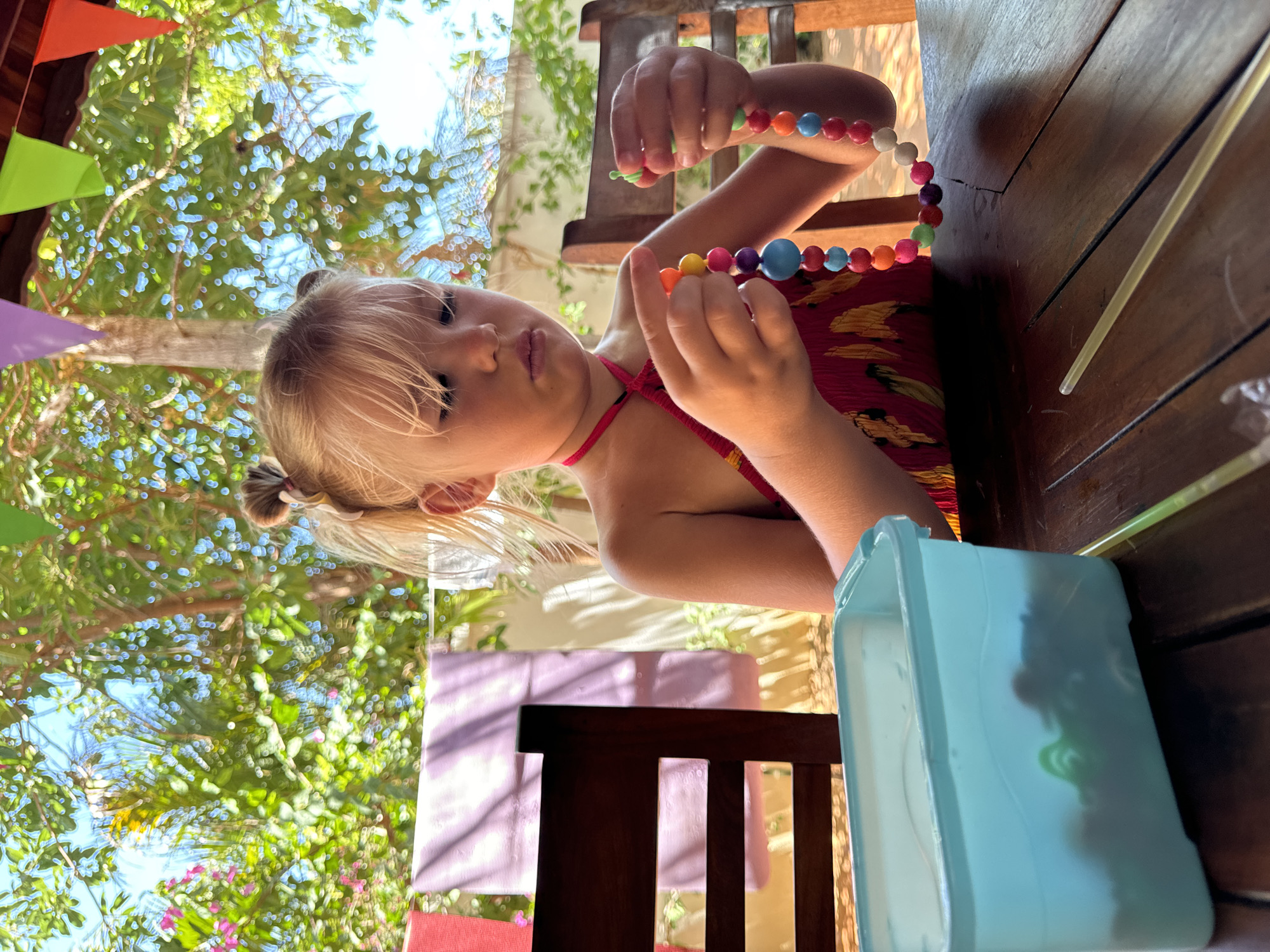 A young blonde girl with pigtails is seated at a wooden table in Mũi Né, Vietnam, holding a colorful bead necklace. She wears a red patterned halter top. A translucent blue container sits beside her on the table. Above, trees and decorative colored pennants are visible.