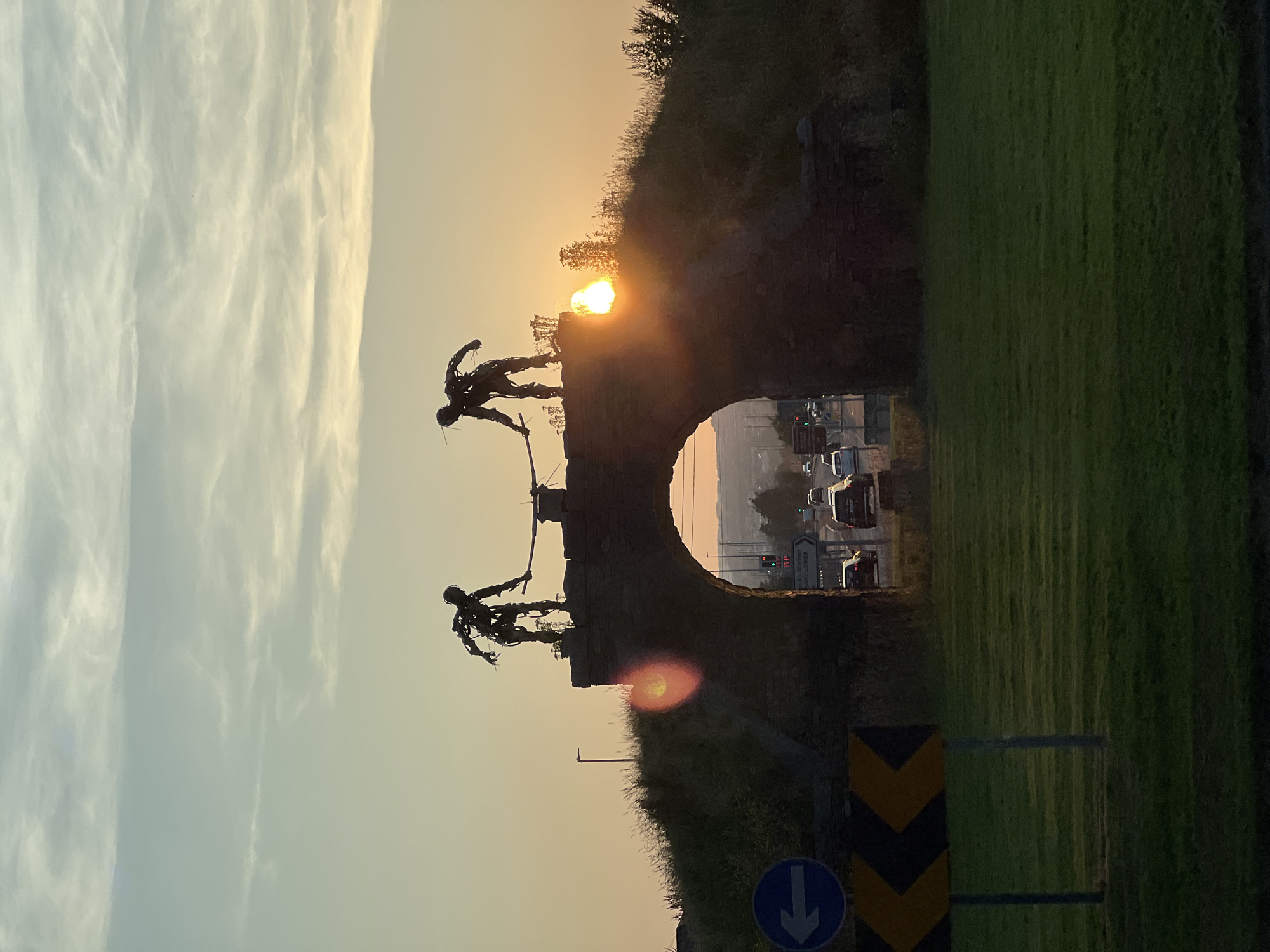 Two metallic, skeletal sculptures of human figures stand atop a stone archway, silhouetted against a sunset sky. The figures appear to be holding a long pole or beam between them. The archway is part of a grassy, raised embankment, through which a road with traffic and traffic lights is visible. To the left of the arch, a blue directional sign with a white arrow pointing left is mounted on a pole, accompanied by a yellow and black chevron road sign indicating a sharp curve. The foreground is a mown grassy area. The overall lighting suggests it is late evening, with the sun setting behind the archway and casting a warm glow. The location is the Oldtown Roundabout in Letterkenny, County Donegal, Ireland, featuring the "Spire of Hope" sculpture.