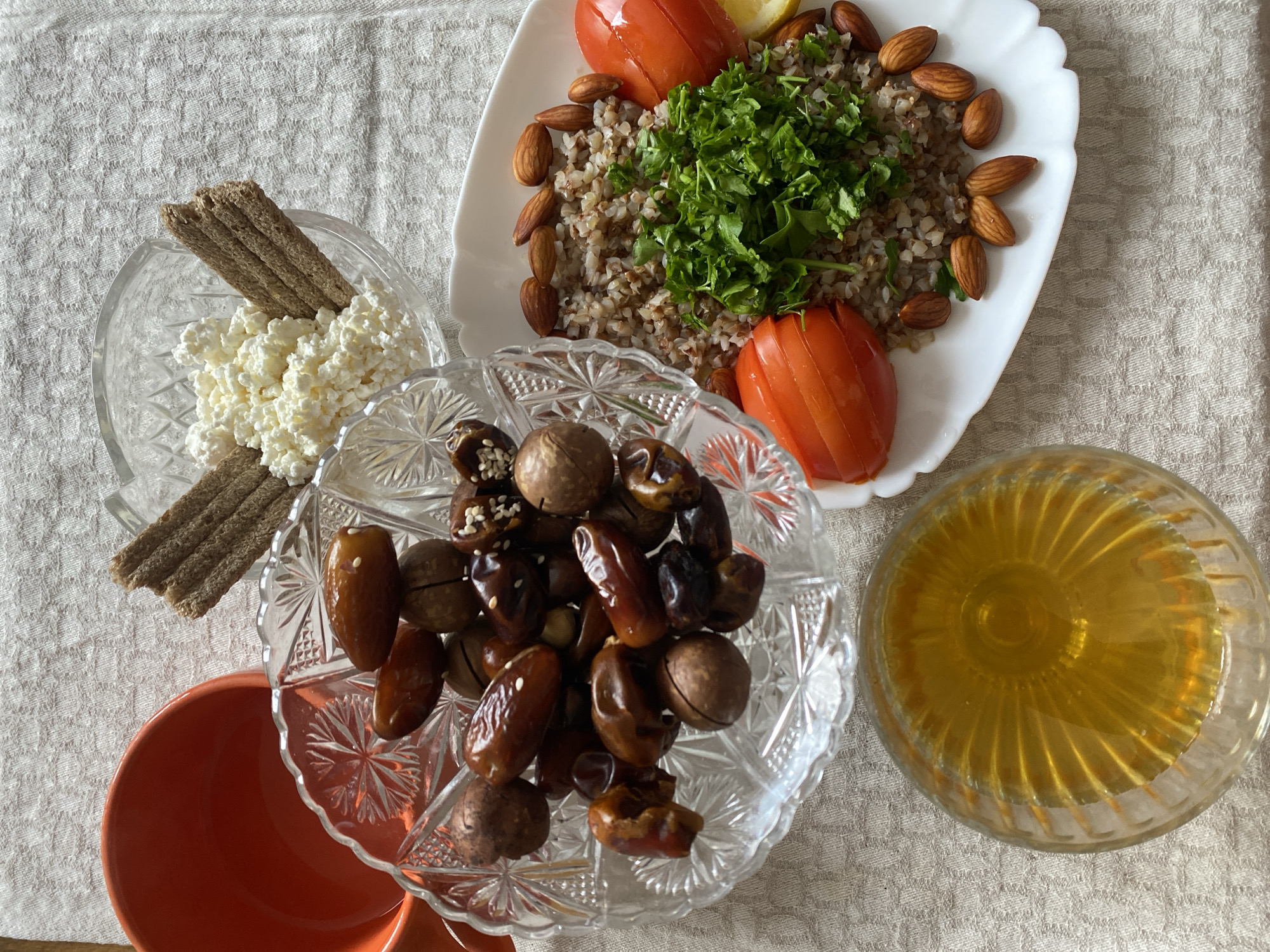 A close-up, overhead shot depicts a breakfast spread on a textured, light-colored tablecloth. In the upper left is an orange ceramic bowl. To its right, a crystal bowl contains white cottage cheese interspersed with small, rectangular pieces of dark bread. Below this, another crystal bowl is filled with dates and macadamia nuts. In the lower left is a glass bowl of golden liquid, likely honey. To the right, a white oval plate holds a pile of cooked buckwheat, garnished with chopped parsley. Sliced tomatoes and almonds surround the buckwheat, with a wedge of lemon visible on the right edge of the plate. The scene does not provide any specific information about the city of Lida, Belarus.
