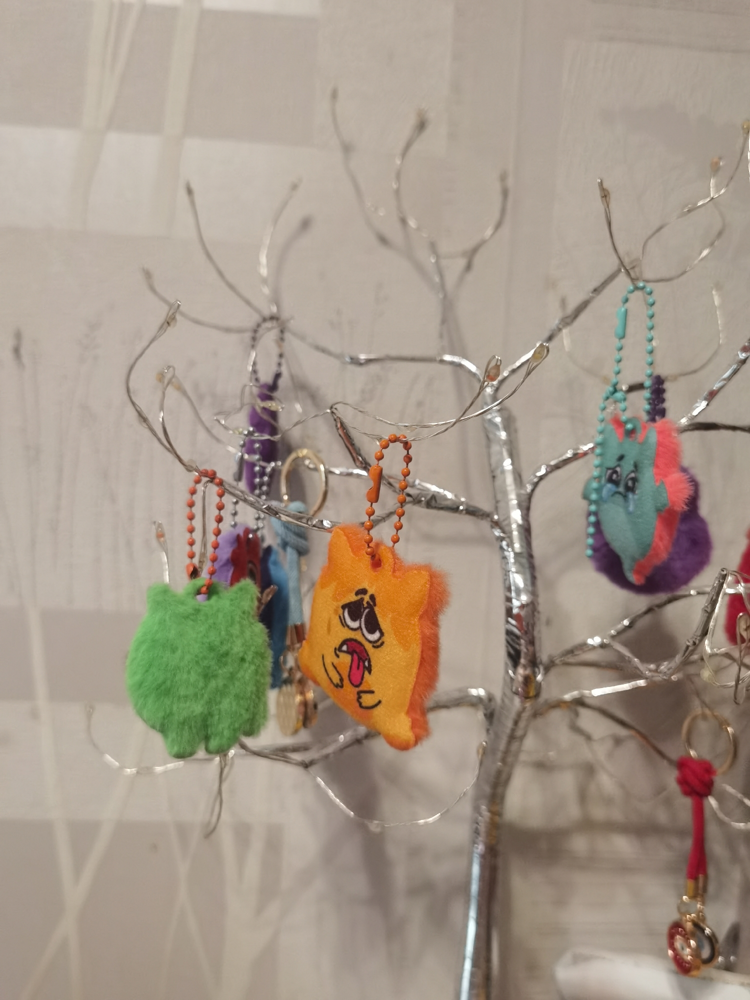 A decorative, metallic, tree-like stand, possibly silver-colored, with numerous thin branches and integrated small, white fairy lights, stands in the foreground, illuminated. Multiple keychains and charms are suspended from various branches. Prominently featured on the left is a fluffy, vibrant green, roughly spherical or blob-shaped charm hanging from a beaded orange chain. To its right, an orange, plush, square-shaped charm with an anthropomorphic face, displaying wide eyes and an open mouth with a protruding red tongue, hangs from a similar orange beaded chain. Further to the right and slightly in the background, a teal or light blue-green plush charm, possibly a character with pink accents, exhibits a sad or crying facial expression with tear-like details beneath its eyes, suspended from a turquoise beaded chain. This teal charm hangs partially over a blurred purple, fluffy object. Other less distinct keychains, including purple and blue fabric elements, metallic rings, and small indiscernible items, are visible among the branches. On the far right, a red corded keychain with a metallic ring and small, ornamental elements hangs. The background consists of a light-toned, possibly off-white or pale gray wall, featuring subtle textural patterns and faint, blurred vertical and horizontal lines, possibly indicating a window or structural elements. The overall scene is brightly lit.