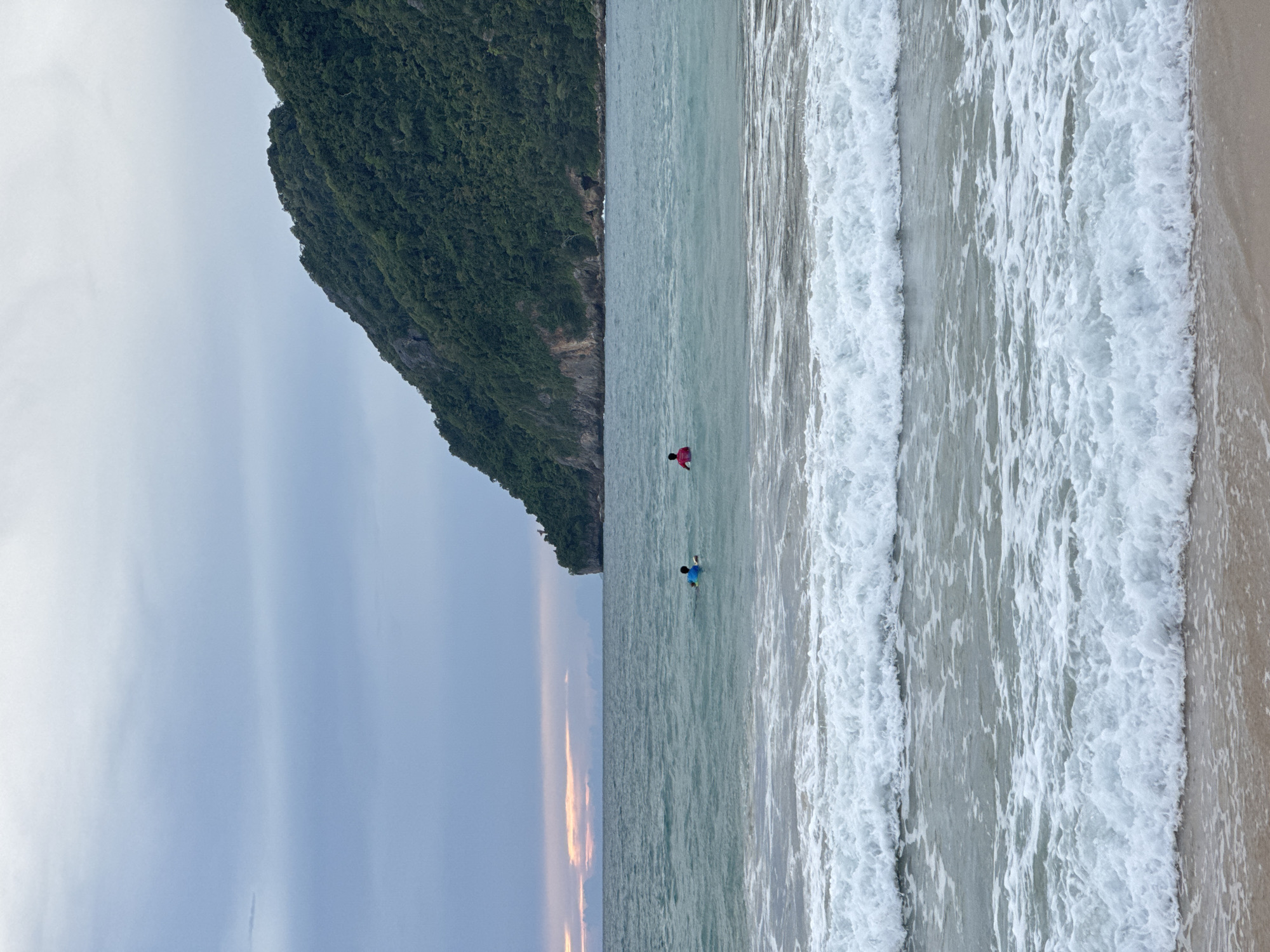 The image depicts a coastal scene with a lush, tree-covered mountain jutting out into the turquoise ocean. The sky is overcast with soft, diffused light. In the foreground, the sandy beach meets the water, where several waves are breaking with white foam. Two individuals are visible in the water; one, wearing a blue top and yellow float, is further out, while another, wearing a red top, is closer to the shore. The scene is set at Meunasahbale, Indonesia.