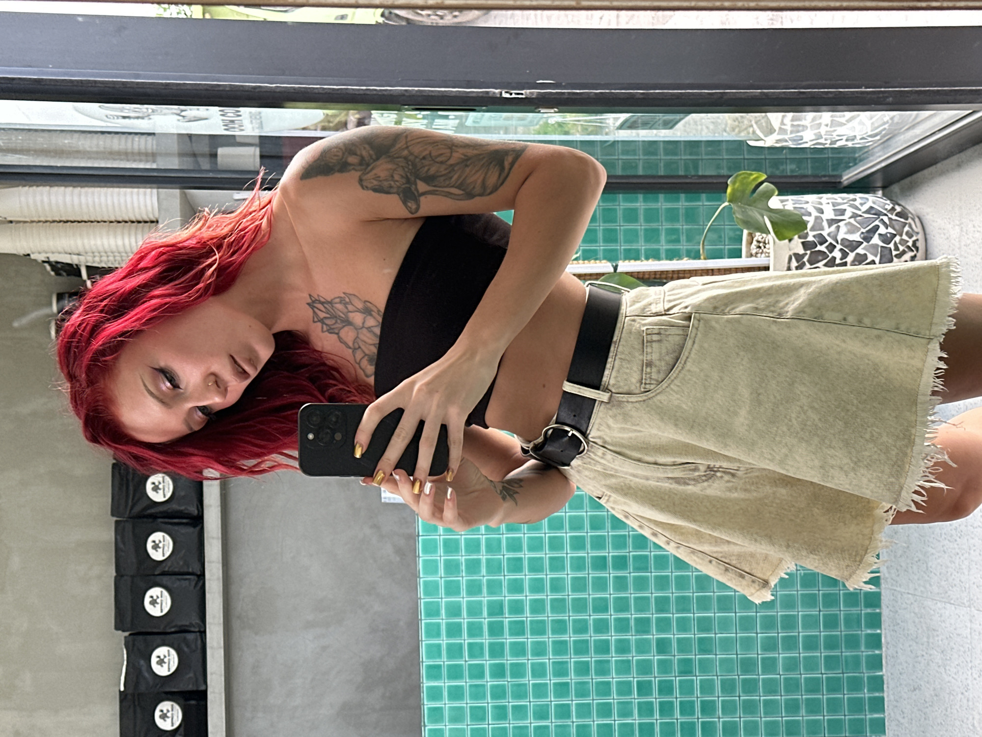 A woman with red hair stands in front of a reflective surface, possibly a mirror, taking a picture of herself with a smartphone. She wears a black tube top, a light-colored denim skirt with frayed edges, and a black belt with a distinctive buckle. Her right arm has a detailed tattoo.
Behind her, the background features a wall covered in small, turquoise tiles. Above this is a shelf with a potted plant. To the left are several black packages stacked vertically. The reflection indicates a cafe setting, possibly located in North Kuta, Indonesia.