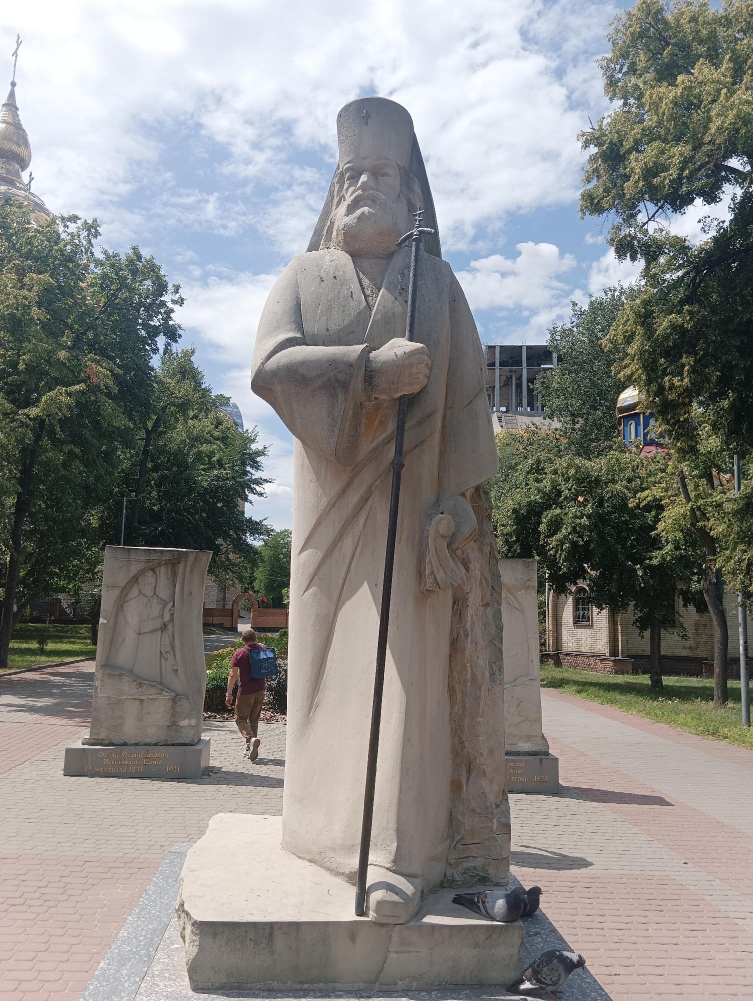 A large stone statue of a clergyman stands prominently in the foreground. He is depicted wearing a tall headdress and a long robe, holding a staff in his right hand. To the left of the main statue, there is another stone monument with a relief carving of a person. A person wearing a maroon shirt and brown pants, with a blue backpack, is walking away from the camera on a paved pathway in the middle ground. The pathway is made of red and grey bricks and is lined with trees on both sides. In the background, there are buildings with golden and blue domes, suggesting a religious or historical site. The sky is partly cloudy with white cumulus clouds against a bright blue background. The sun is shining, casting shadows on the pathway. Two pigeons are visible near the base of the main statue, one is sitting on the statue's base and the other is on the pathway.
Visible text on the monument to the left of the main statue reads: "Феликса Станиславовича Лещенка-Полтавца. 19 августа 1841 - 1931".
Visible text on the monument on the right reads: "Они растут из земли..." followed by "26 серпня 1928".